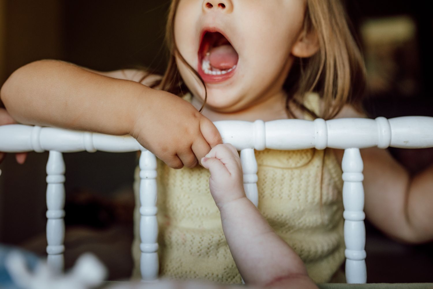 A small child in a yellow sweater yawns while gripping the white spindles of a bannister or railing.