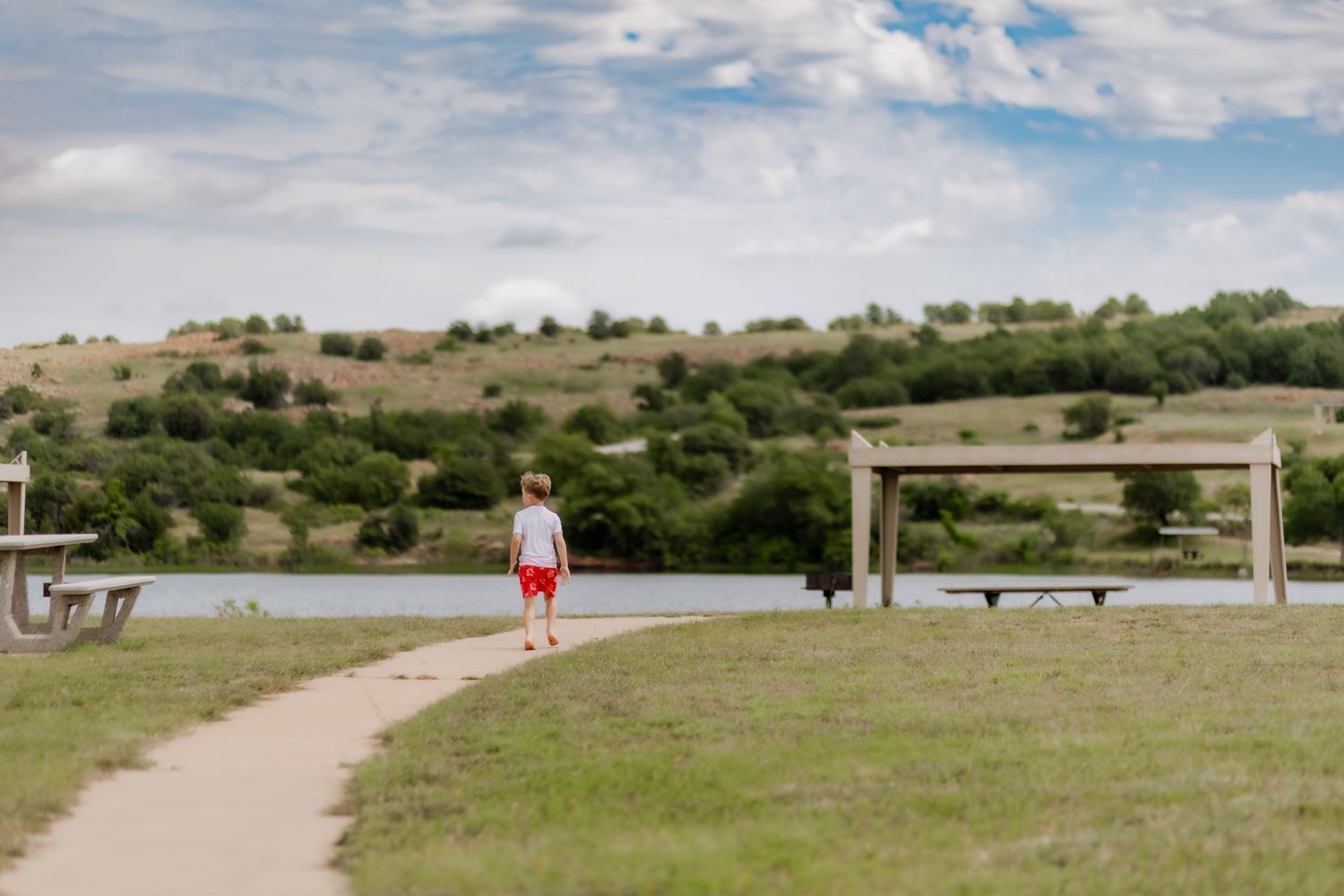Person walking on dirt path through grassy landscape with wooden structure in background.