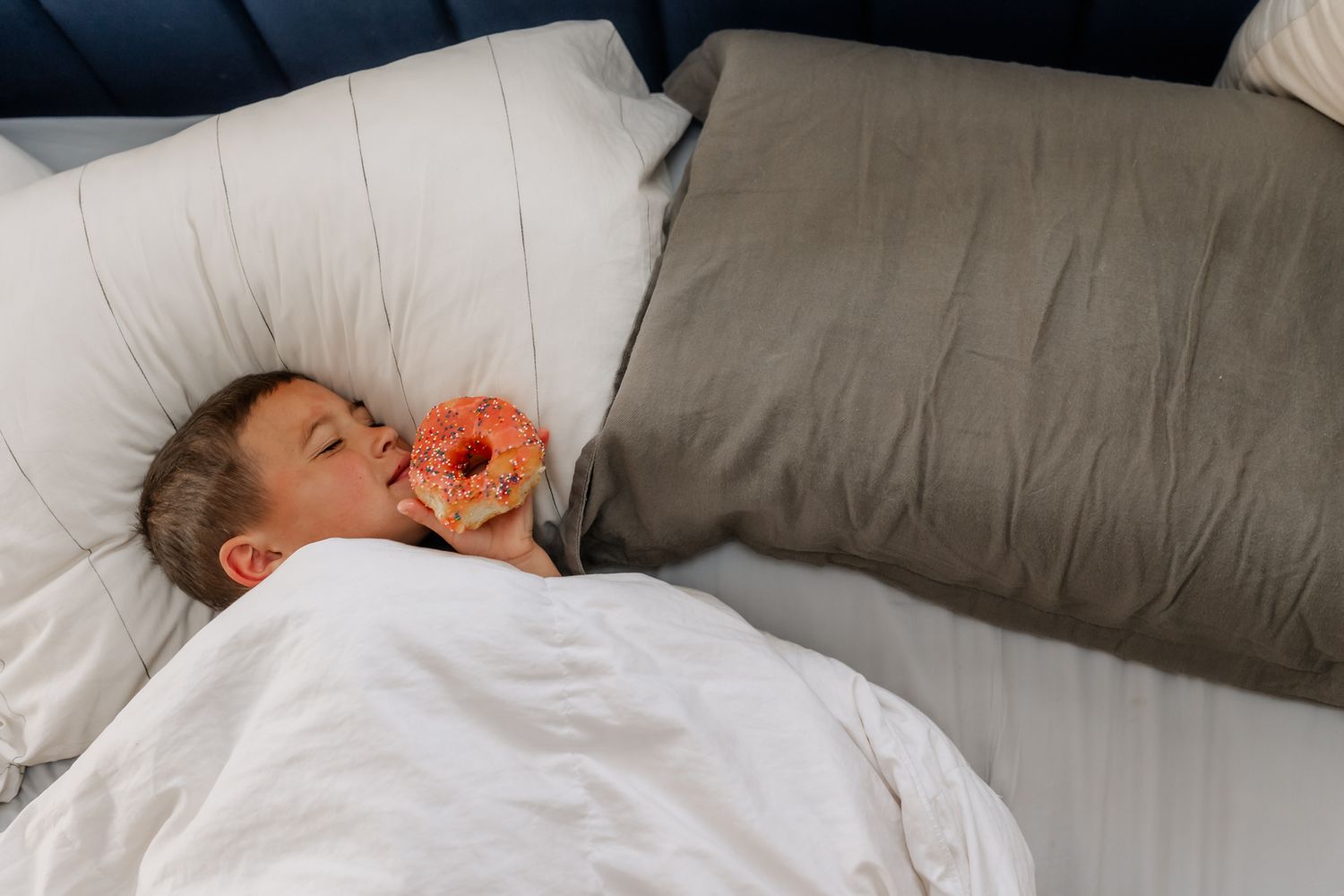 A sleeping child nestles in white bedding while holding an orange-pink donut pillow against their face.
