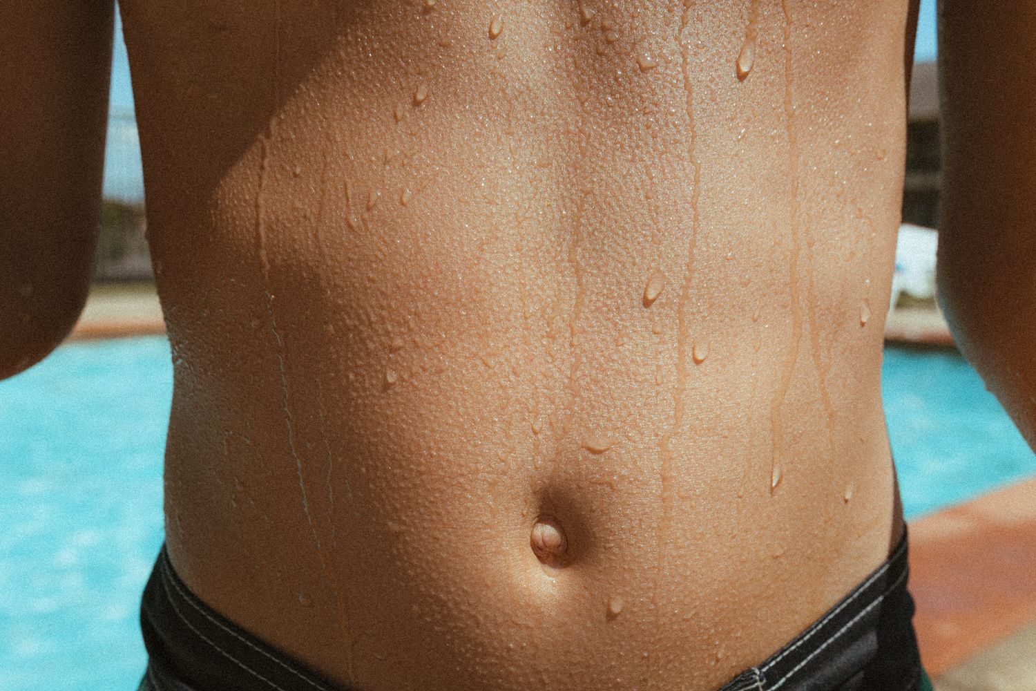 Close-up of a toned wet torso with water droplets near a swimming pool on a sunny day.