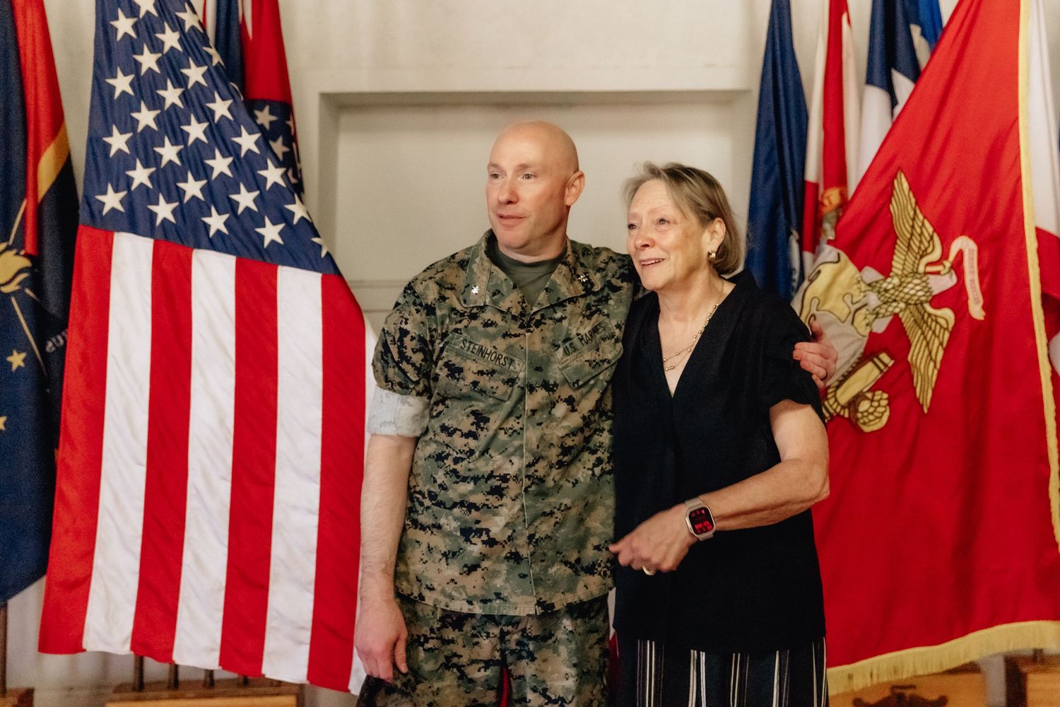 Military serviceman in camouflage uniform poses with elderly woman in front of American and Marine Corps flags.