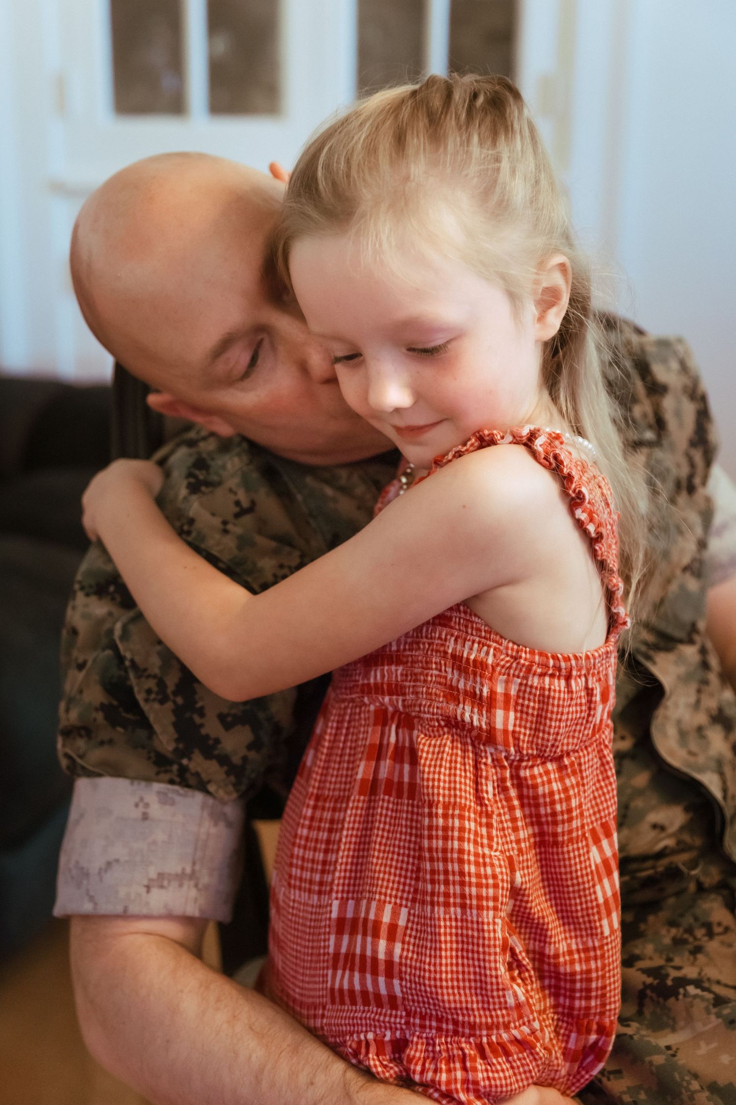 Military father in camouflage uniform hugging his young blonde daughter in a red dress indoors.