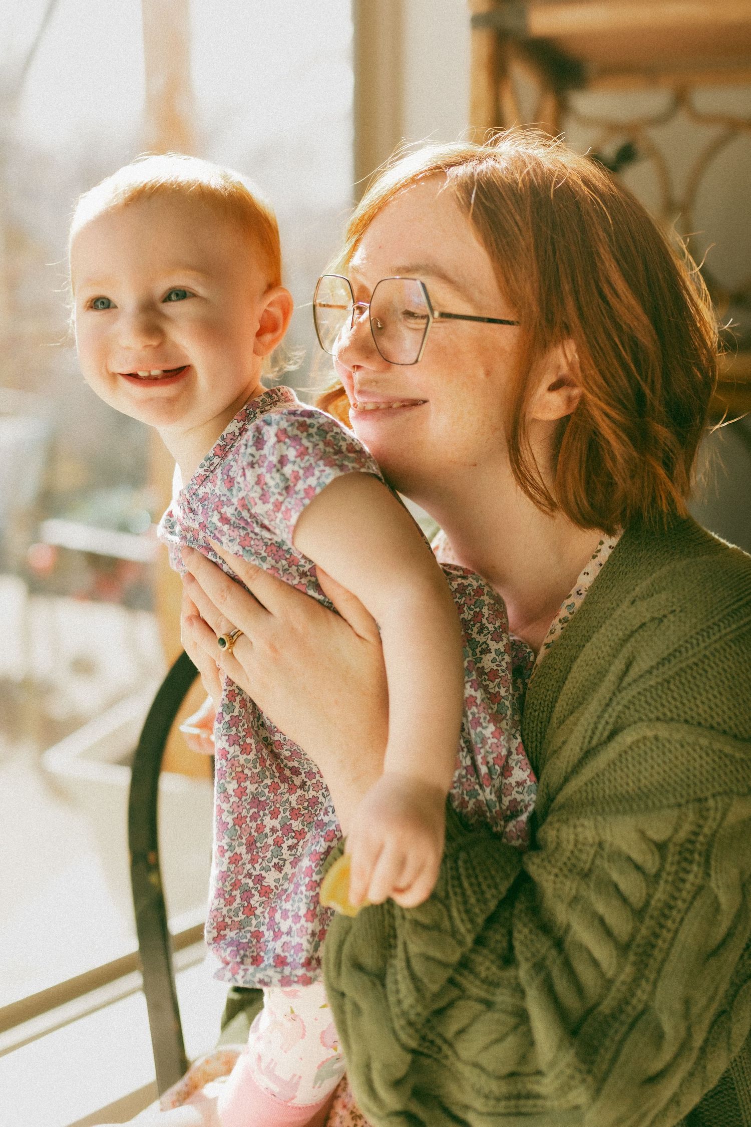 A tender moment captured in warm sunlight as a baby in floral dress smiles while being held near a window.