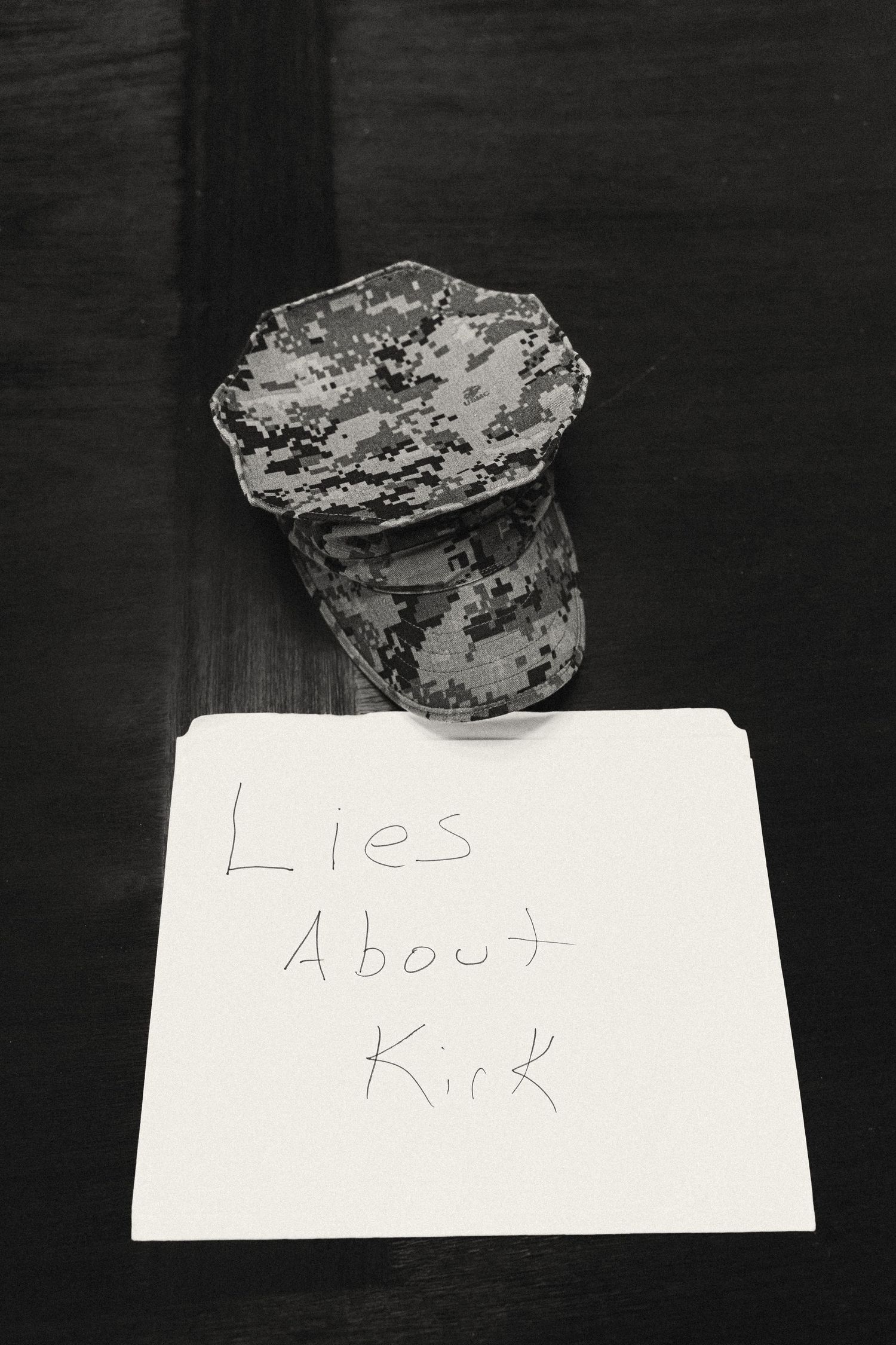 Black and white photo of a military camouflage cap next to a handwritten note reading Lies About Kirk.