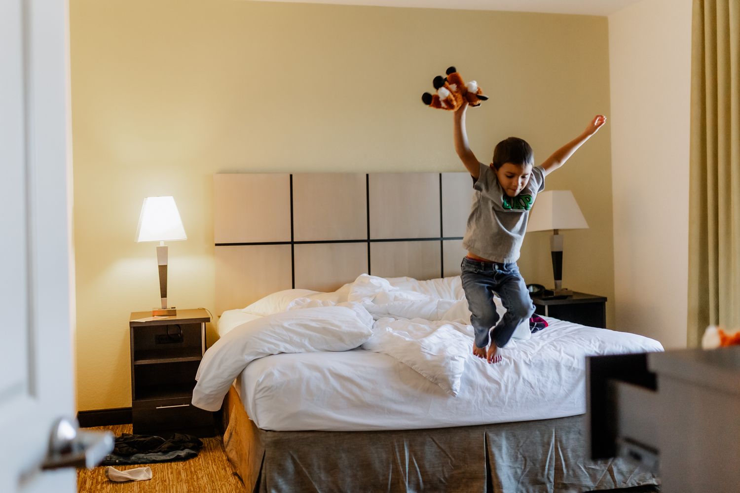 Someone wearing a gray shirt and dark pants jumps energetically on a hotel room bed next to a table lamp.
