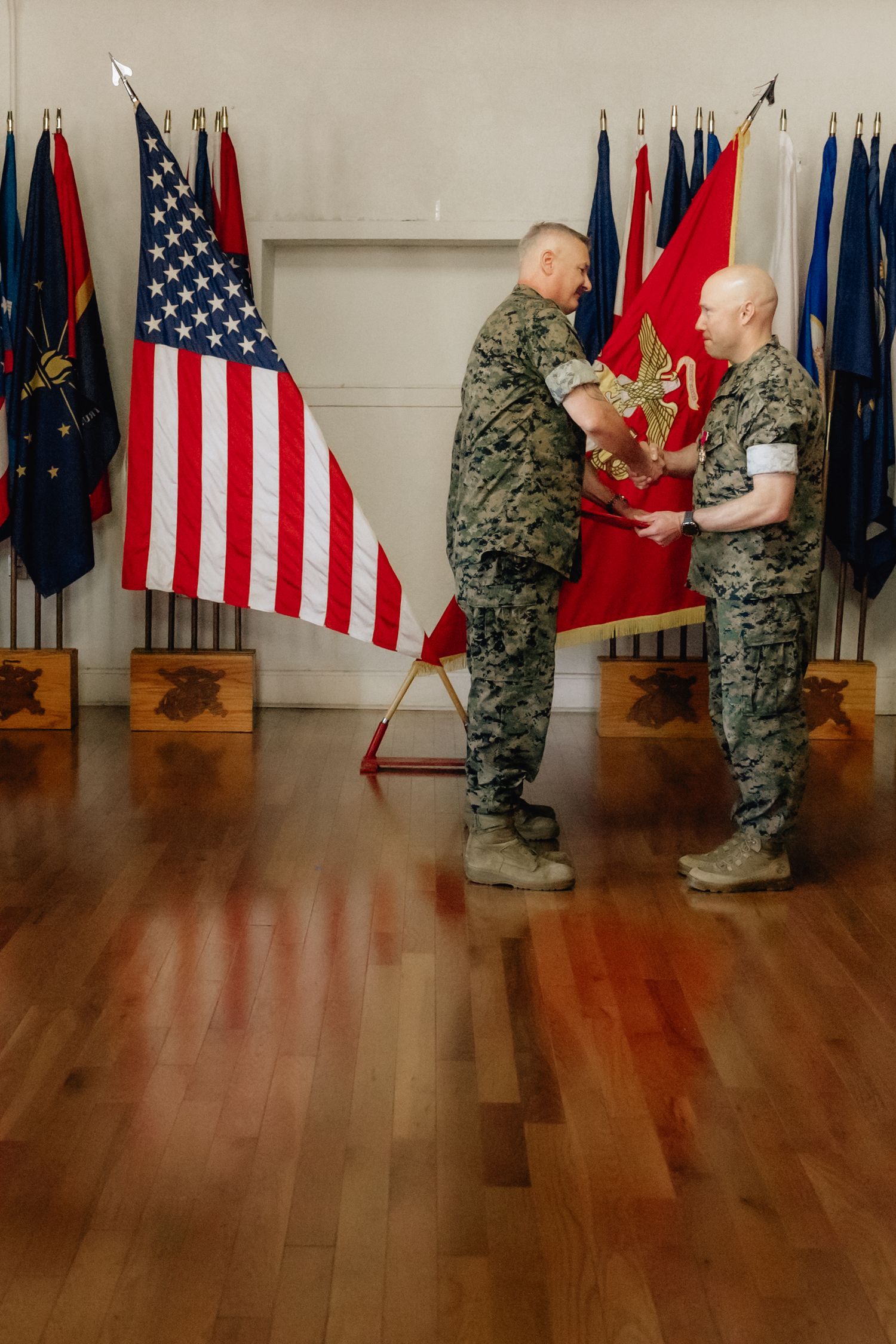 Two Marines in camouflage uniforms exchange a red military flag during a formal ceremony with US flags displayed.