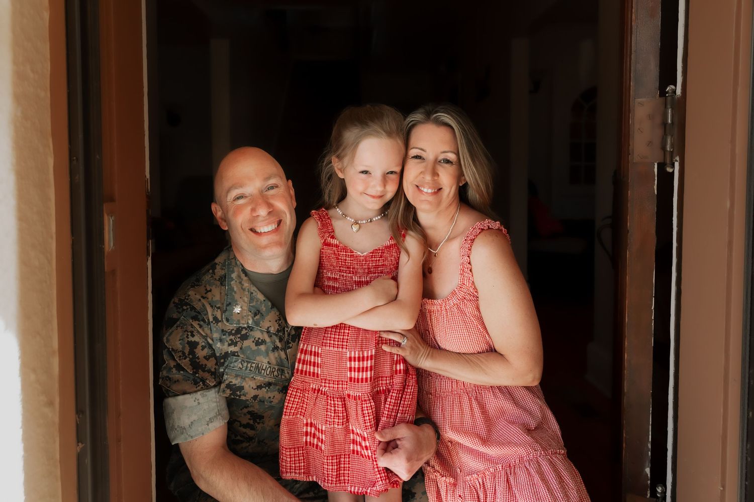 Happy family of three smiling in a doorway, man in military uniform with woman and young girl in red dresses.