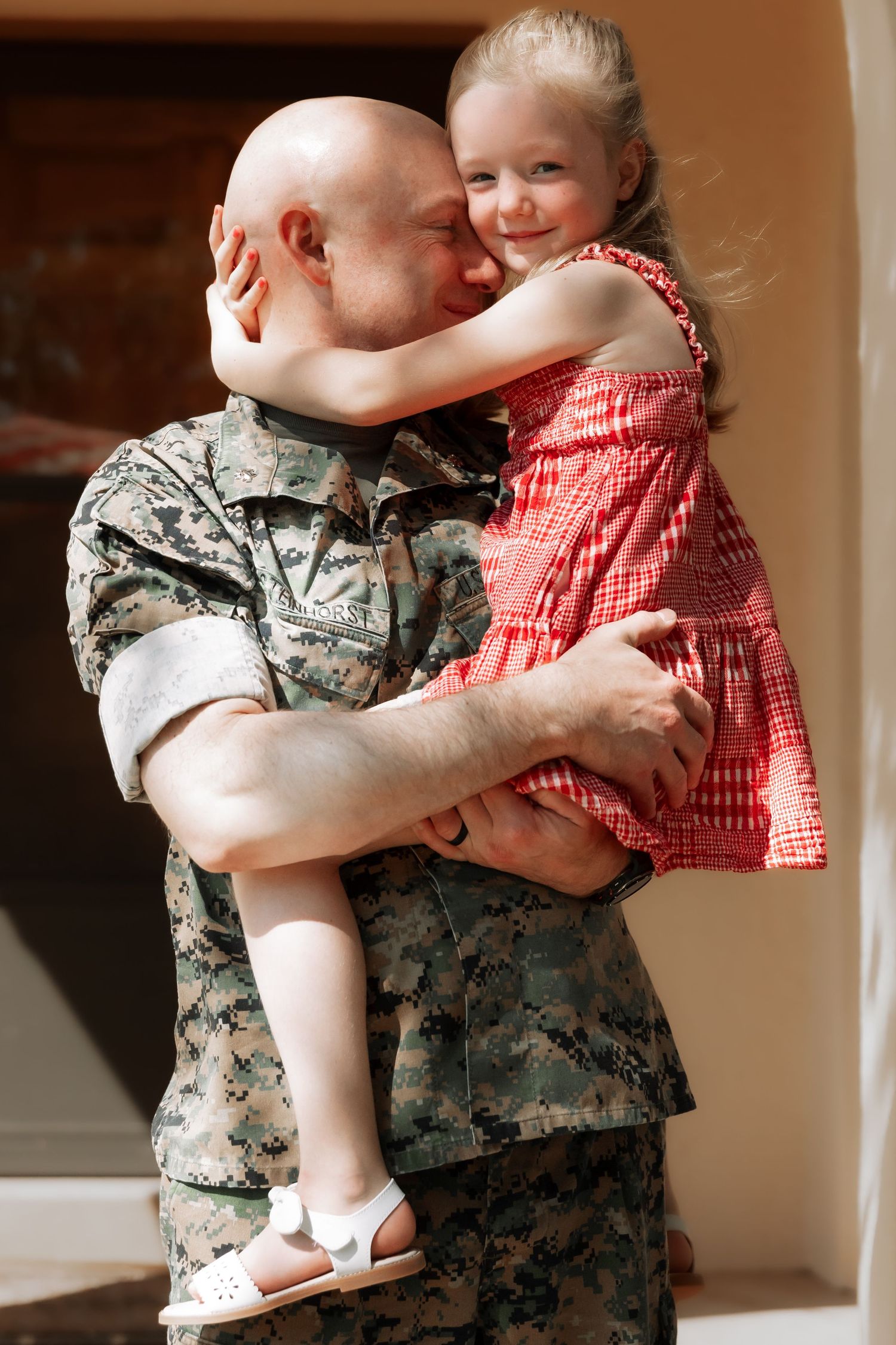 A bald military soldier in camouflage uniform holds his smiling young daughter in a red dress during a homecoming reunion.