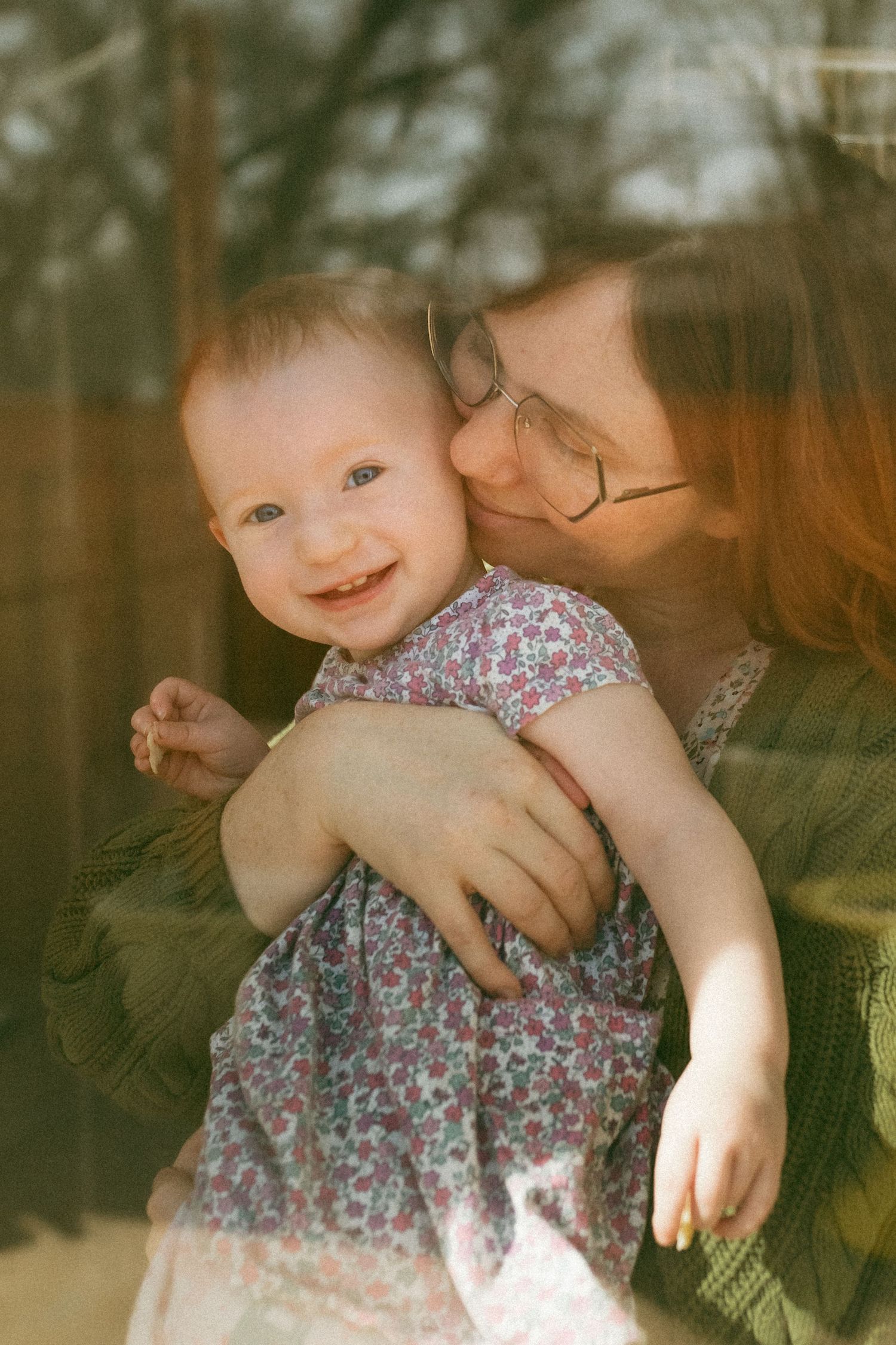 A baby in a floral dress is held lovingly in warm sunlight against a forest backdrop.