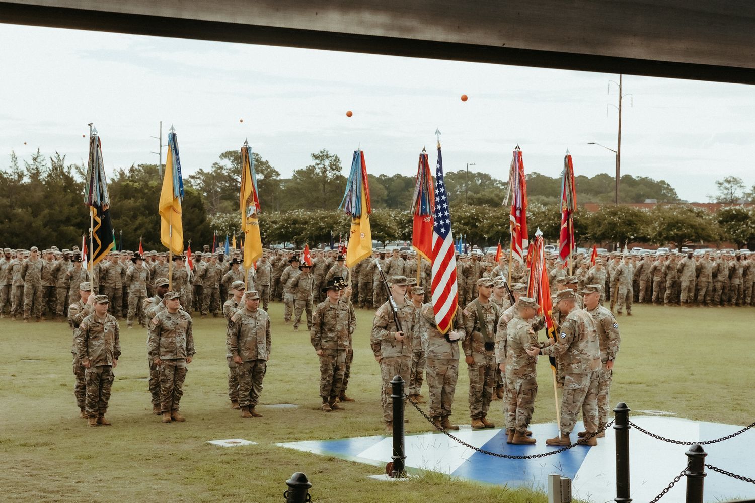 Military personnel stand in formation on a parade ground with flags and banners displayed during a ceremony.