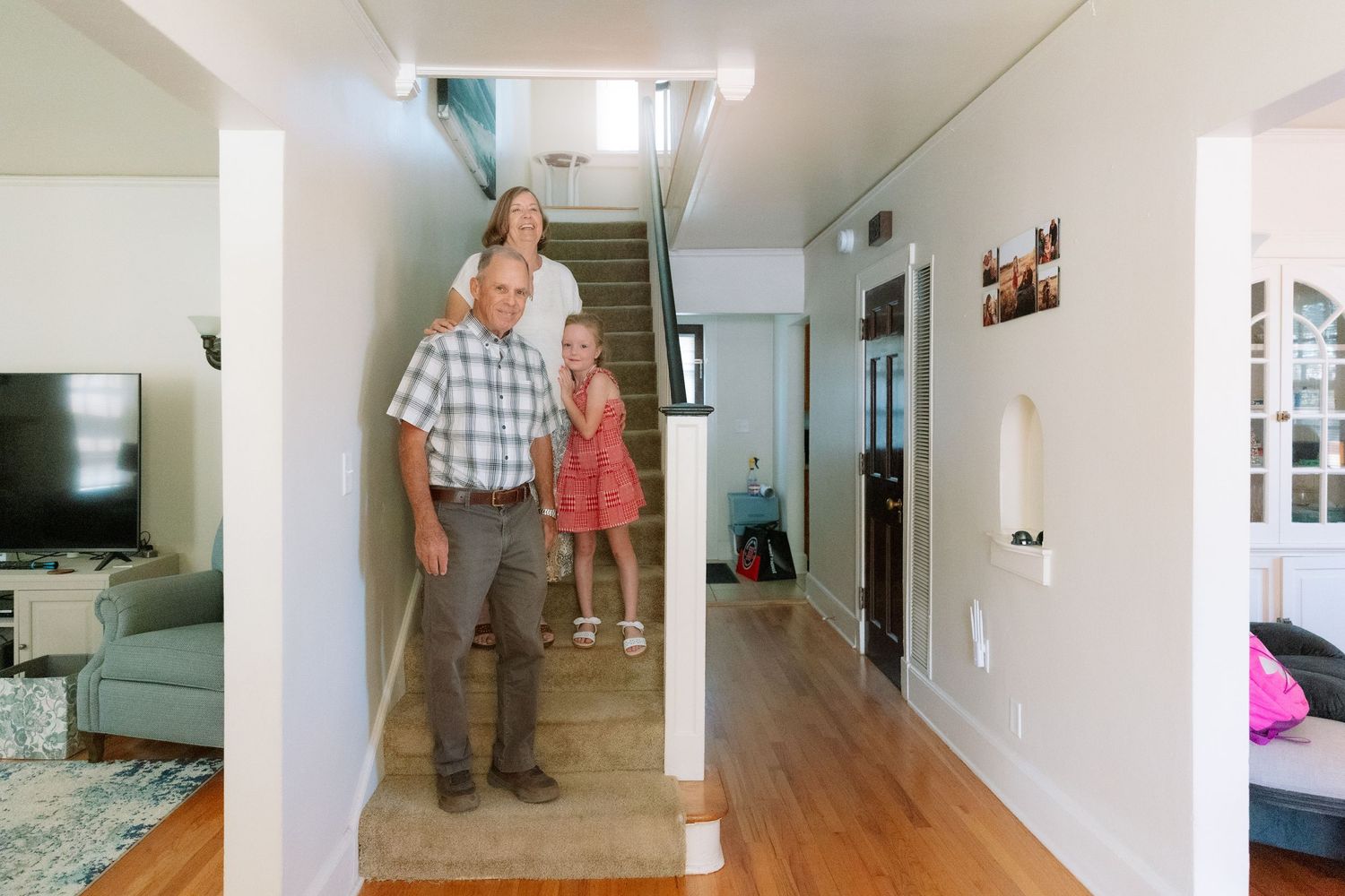 Family of three standing in a bright home hallway with hardwood floors, staircase, and open living room.