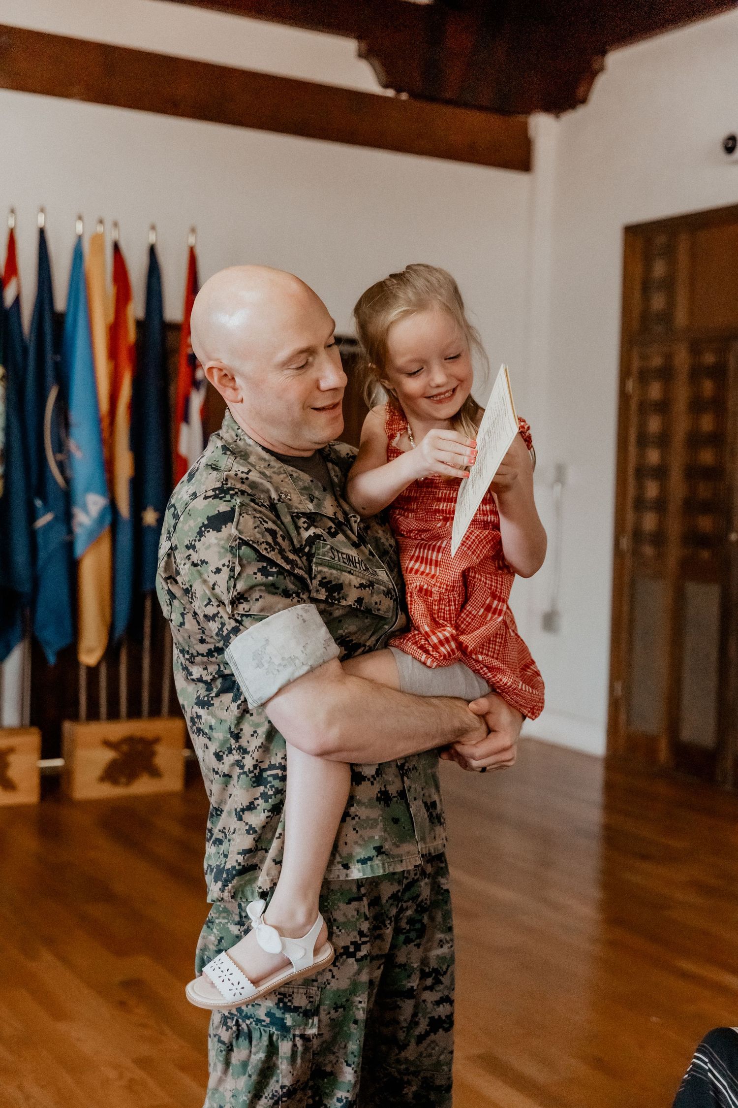 Marine soldier in camouflage uniform holding young blonde girl in red dress, smiling together in flag-decorated room.