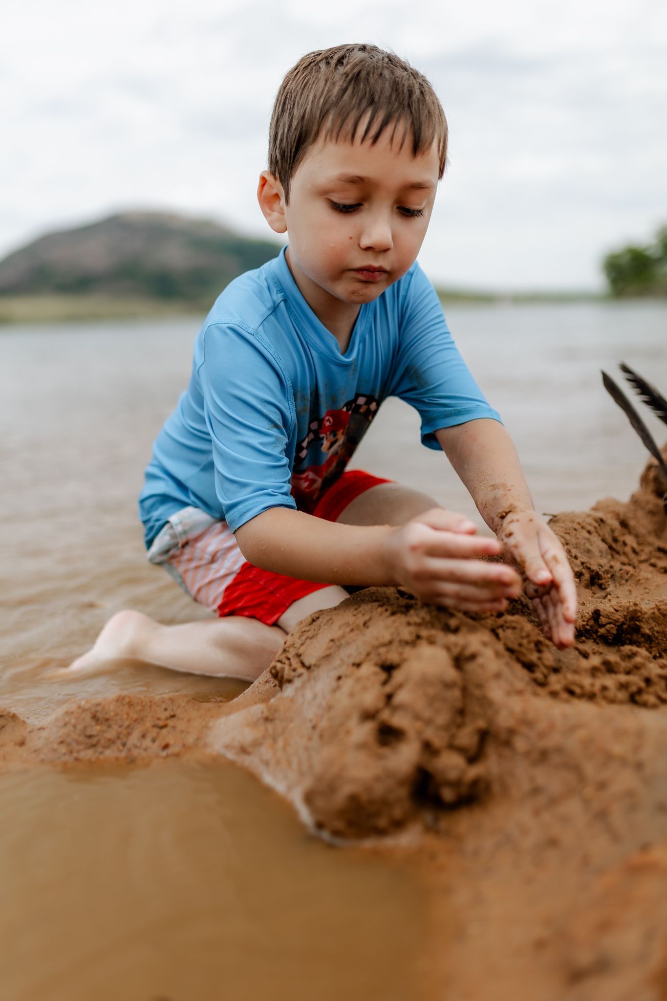 Child in blue shirt playing with wet sand at the edge of a lake on a cloudy day.