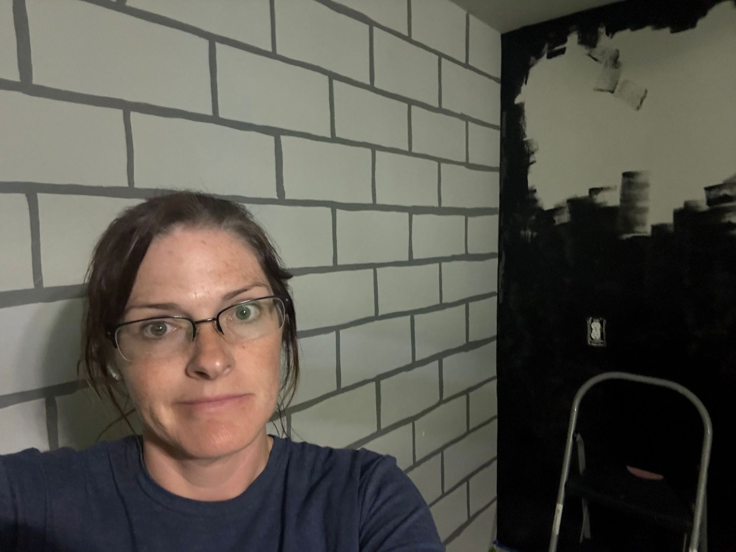 A person in glasses makes an expressive face while posing against a white brick wall background. In-Home Photography Liz Perez