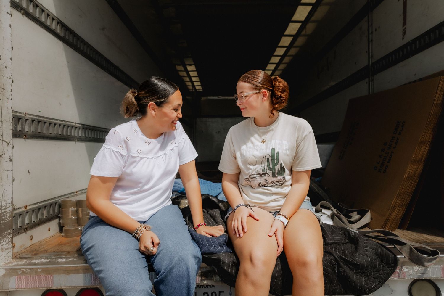 Two women sitting and chatting inside a truck bed, wearing casual white t-shirts in a candid moment.
