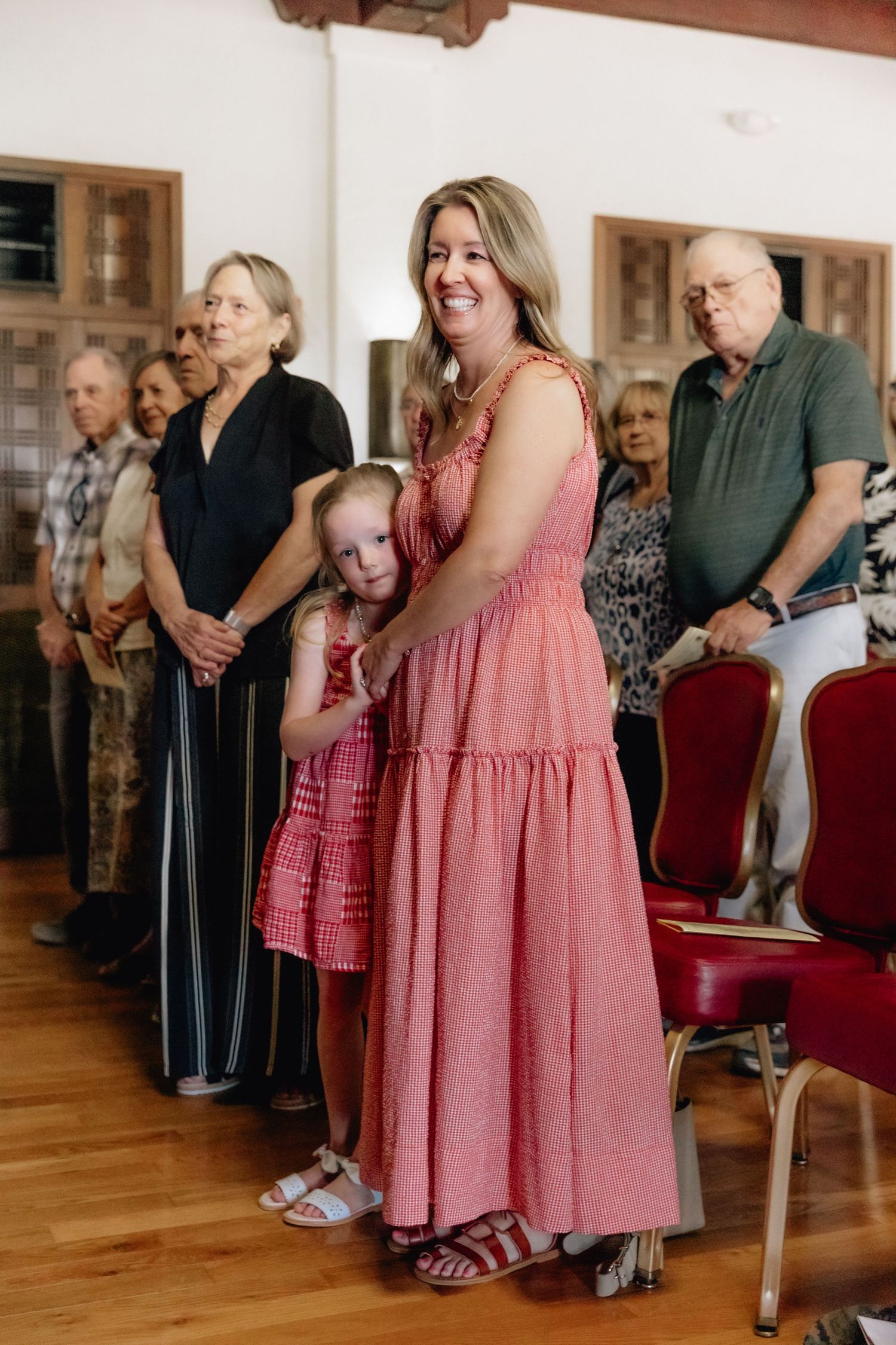 Woman in pink maxi dress smiles while hugging young girl in red dress during a military retirement ceremony.