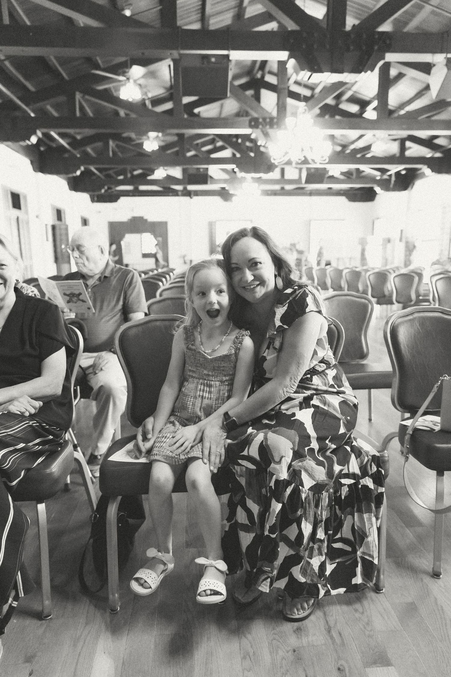 Black and white photo of a woman and young girl sitting together smiling in chairs inside a hall.