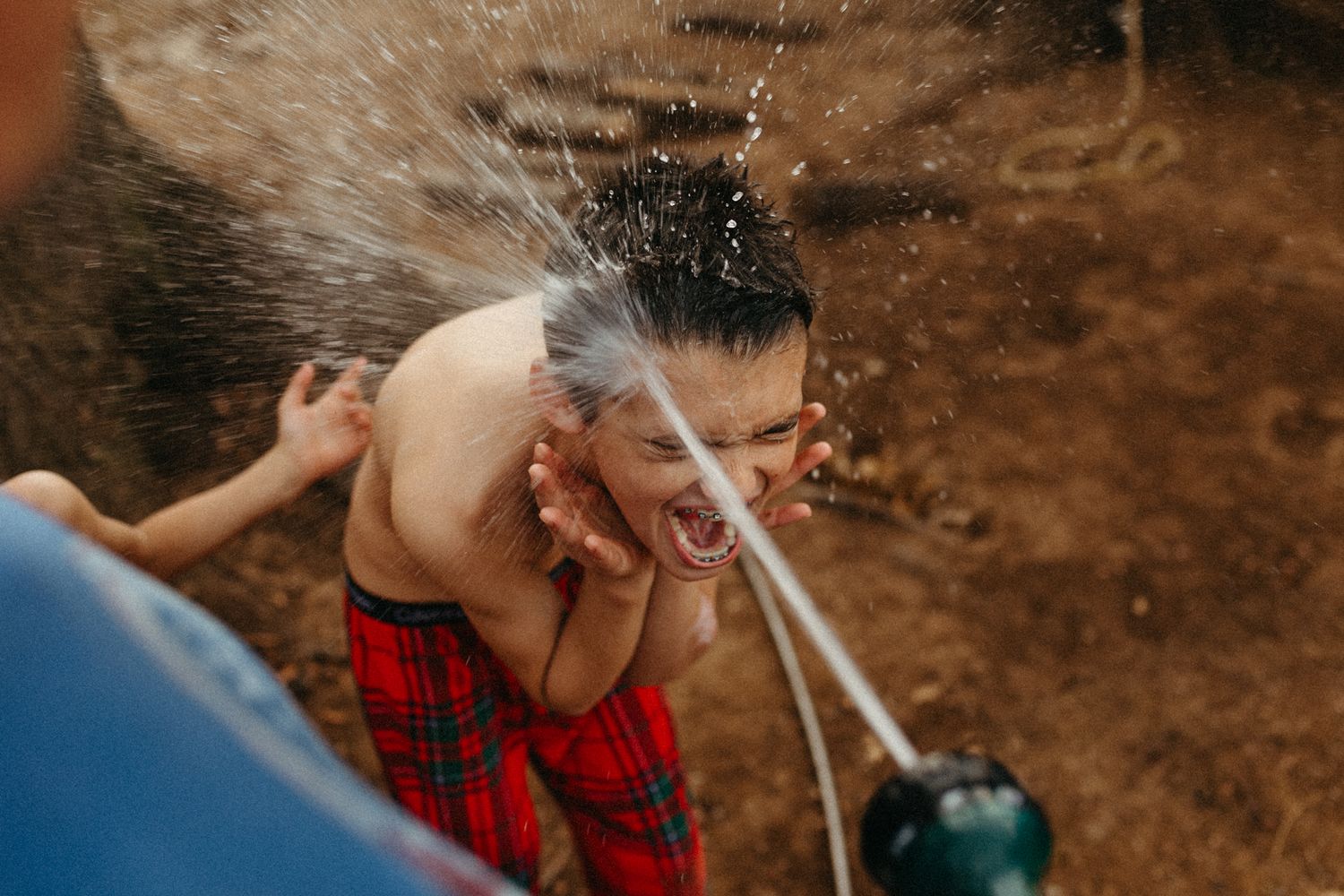 Shirtless boy laughing as water from a hose splashes over his face outdoors.