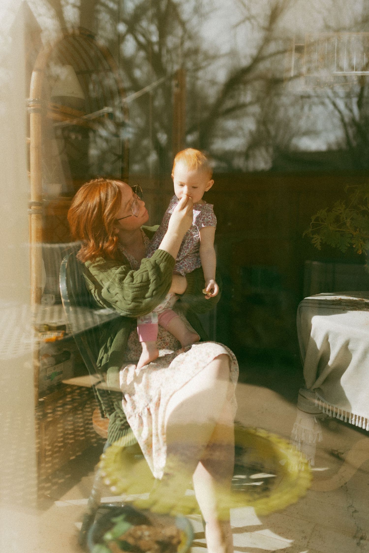A red-haired woman in a green jacket holds a blonde toddler on her lap, photographed through a sunlit window.