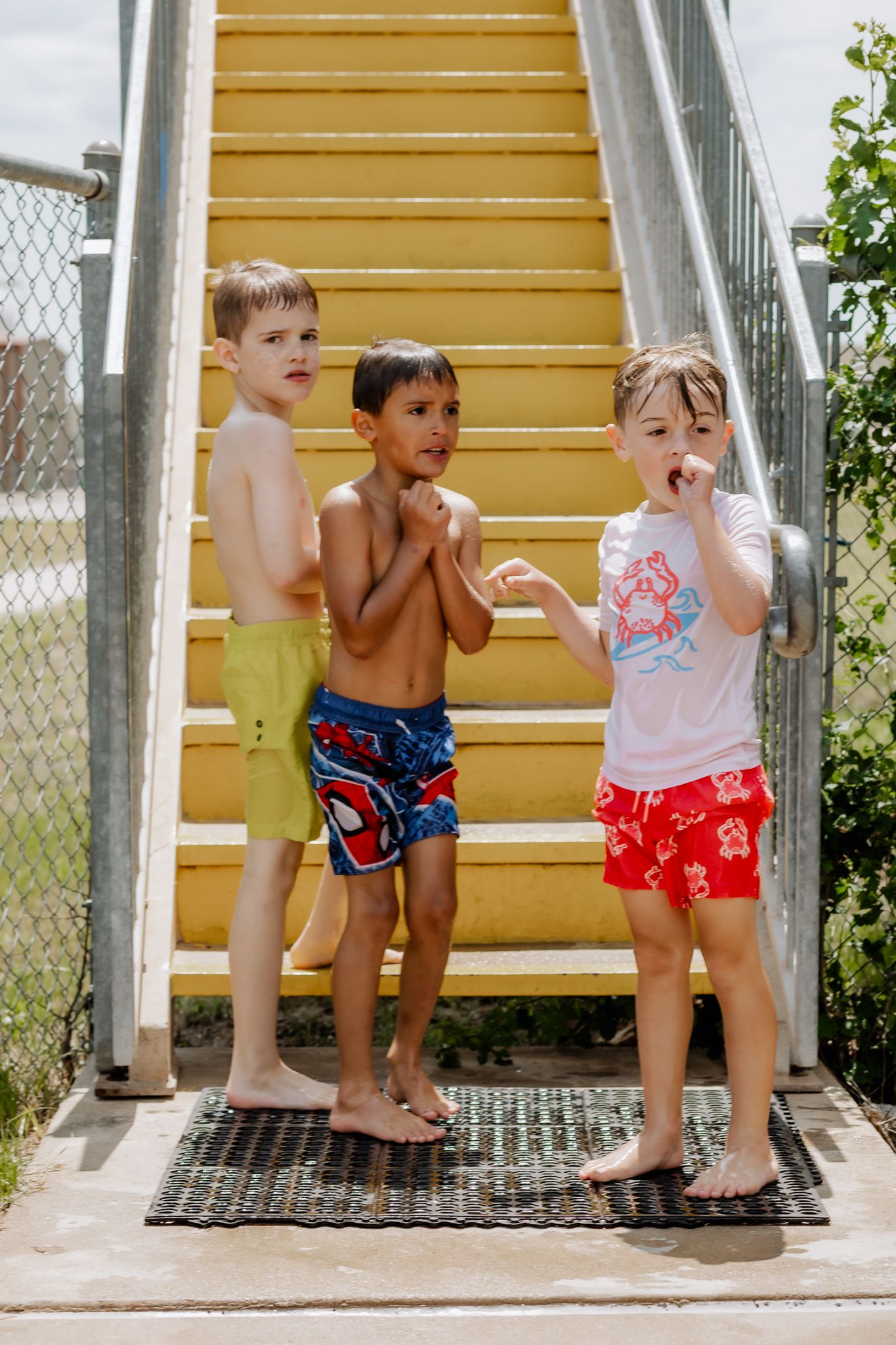 Three small children in swimwear standing together on metal steps at a pool.