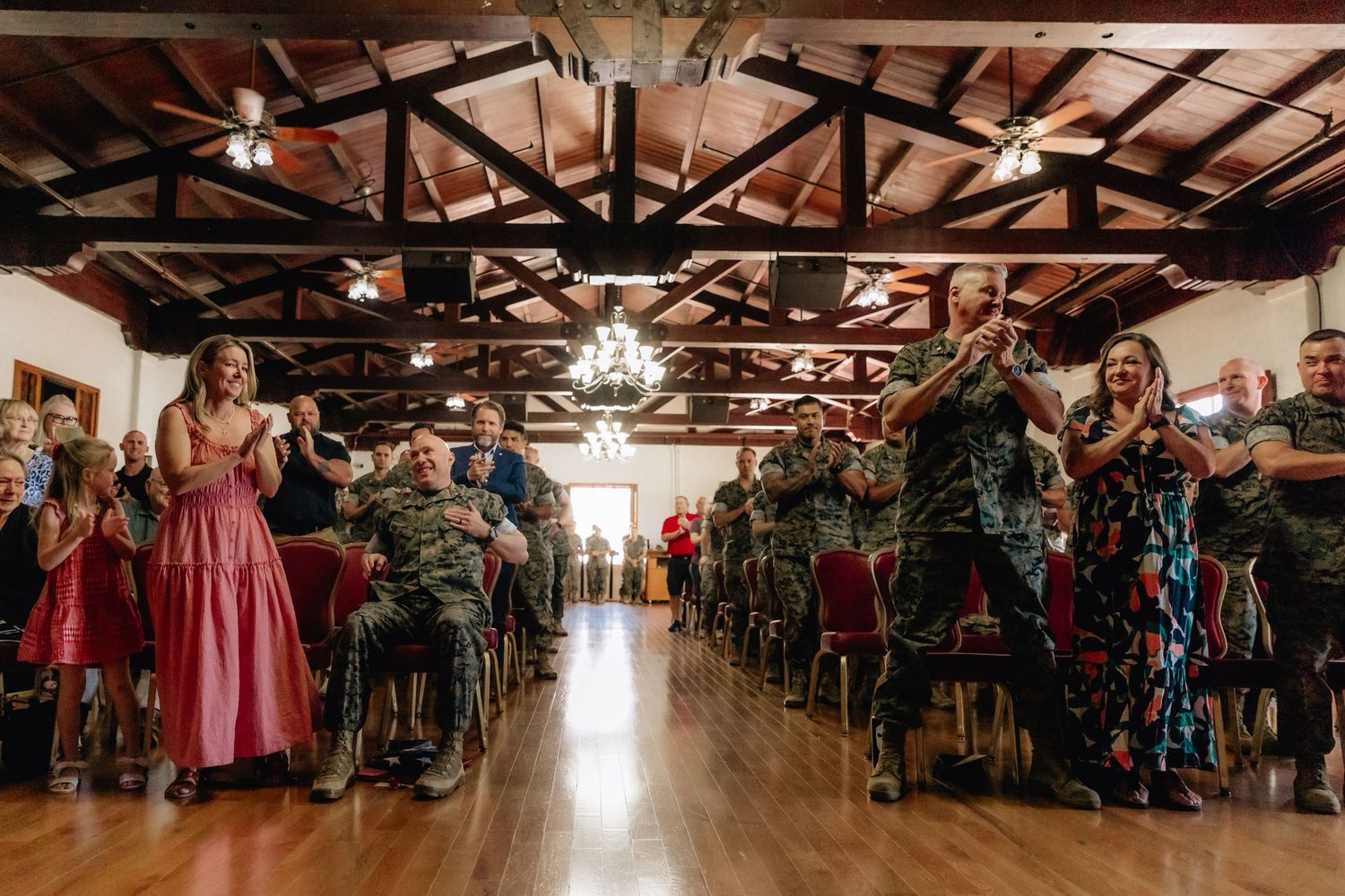 Military wedding reception with guests in camouflage uniforms celebrating in a rustic wood-beamed venue with chandelier.