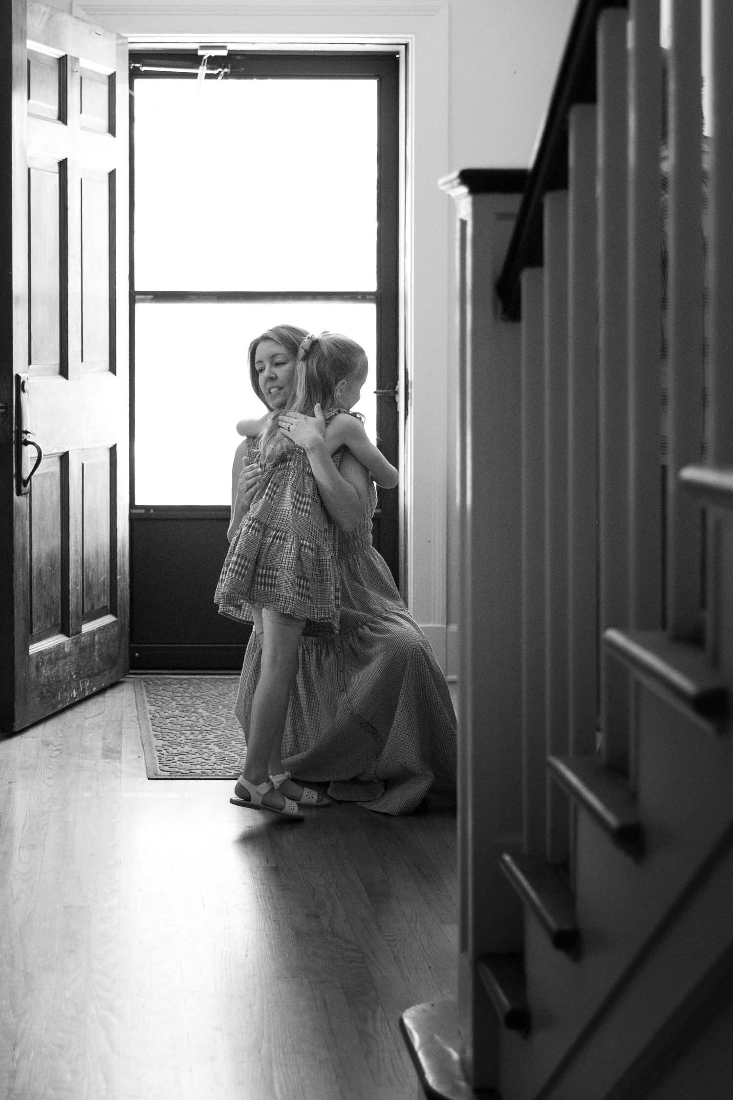 Black and white photo of a woman hugging a young girl in a sunlit home entryway.