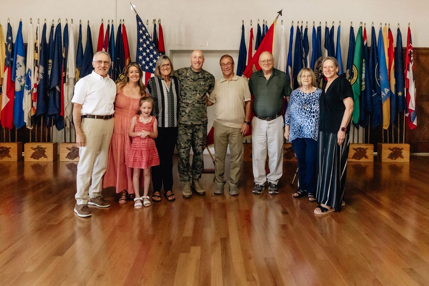 Group of people posing in front of colorful military flags on a wooden floor at a formal ceremony.