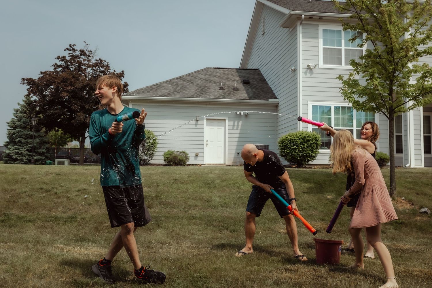 Three people playing with water guns and buckets in a backyard water fight.