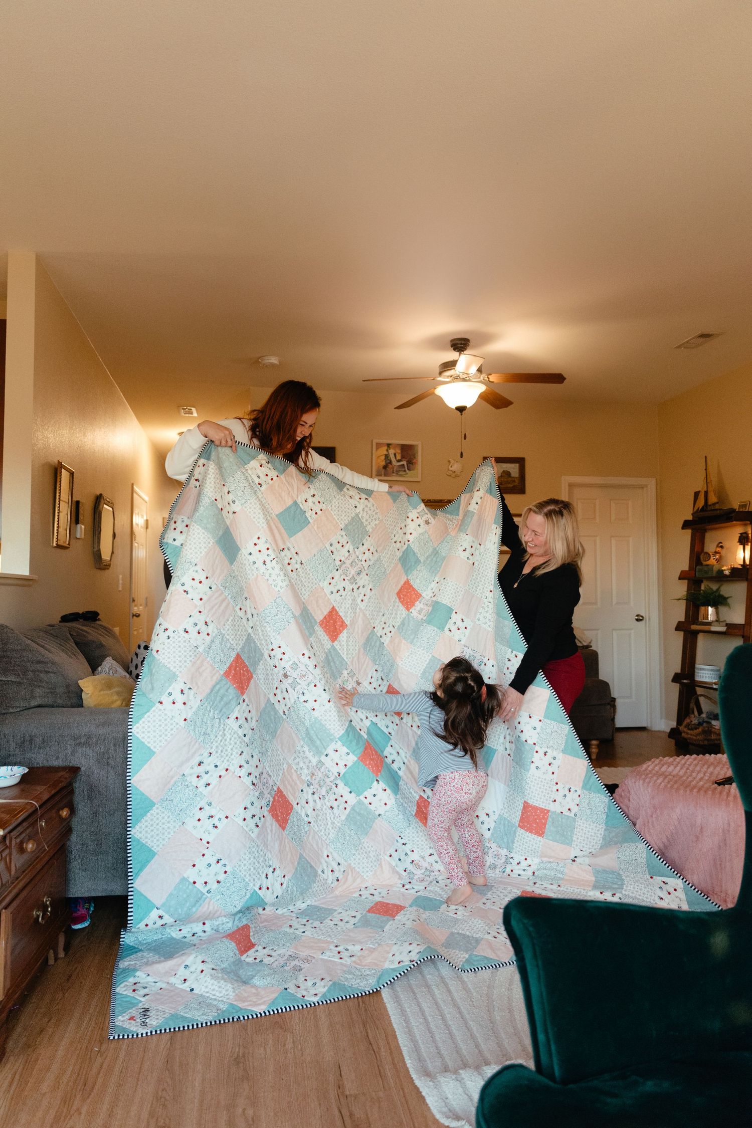 Two women and a child hold up a pastel patchwork quilt in a cozy living room.