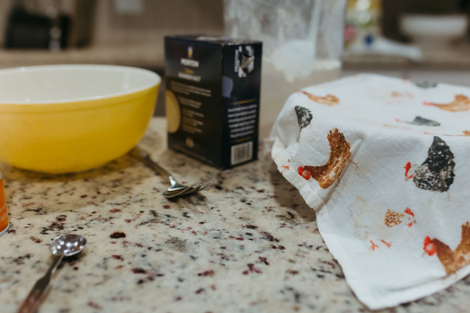 Kitchen counter with yellow mixing bowl, measuring spoons, recipe box, and chicken-print dish towel on granite surface representing mom.