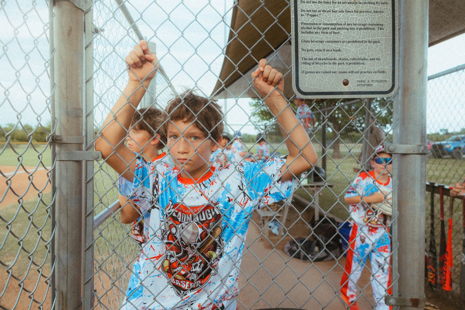 Young baseball player in colorful uniform gripping chain-link fence dugout gate at youth baseball field.
