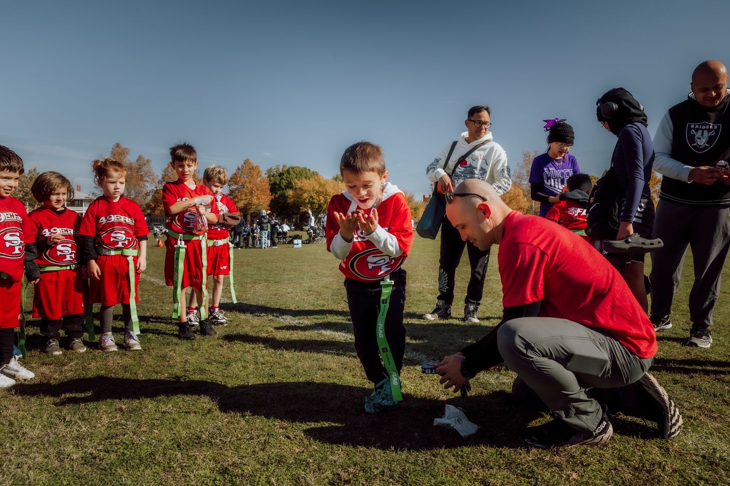 Youth sports team huddles on a grass field during a sunny day wearing red uniforms.