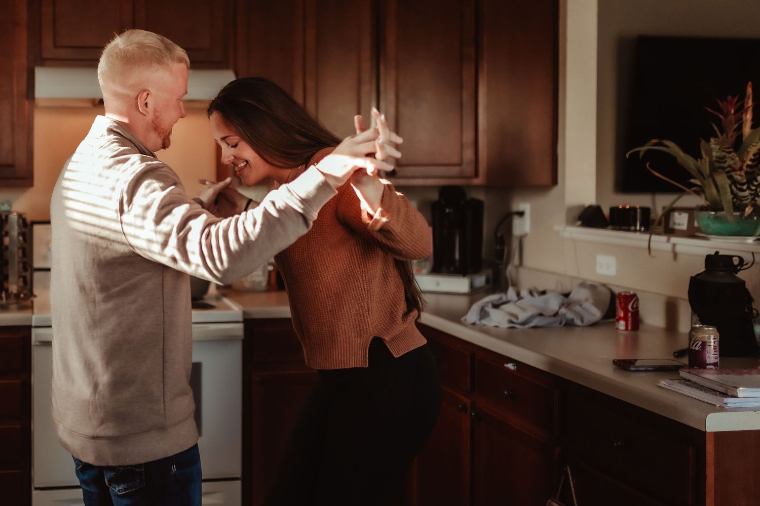 Two people dancing together in a cozy kitchen with warm lighting and wood cabinets.