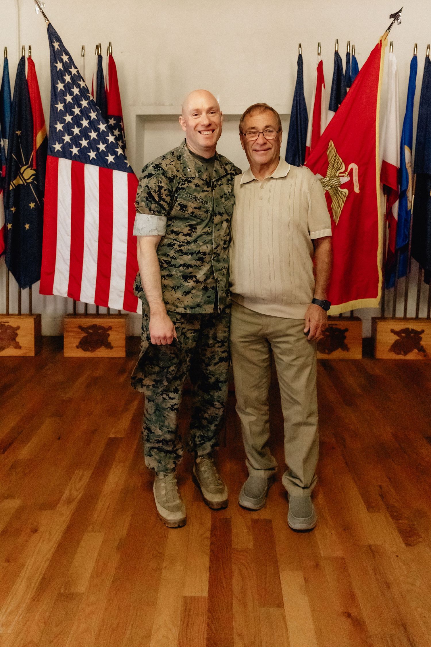 A US Marine in camouflage uniform poses with a civilian man in front of American and military flags on a wood floor.