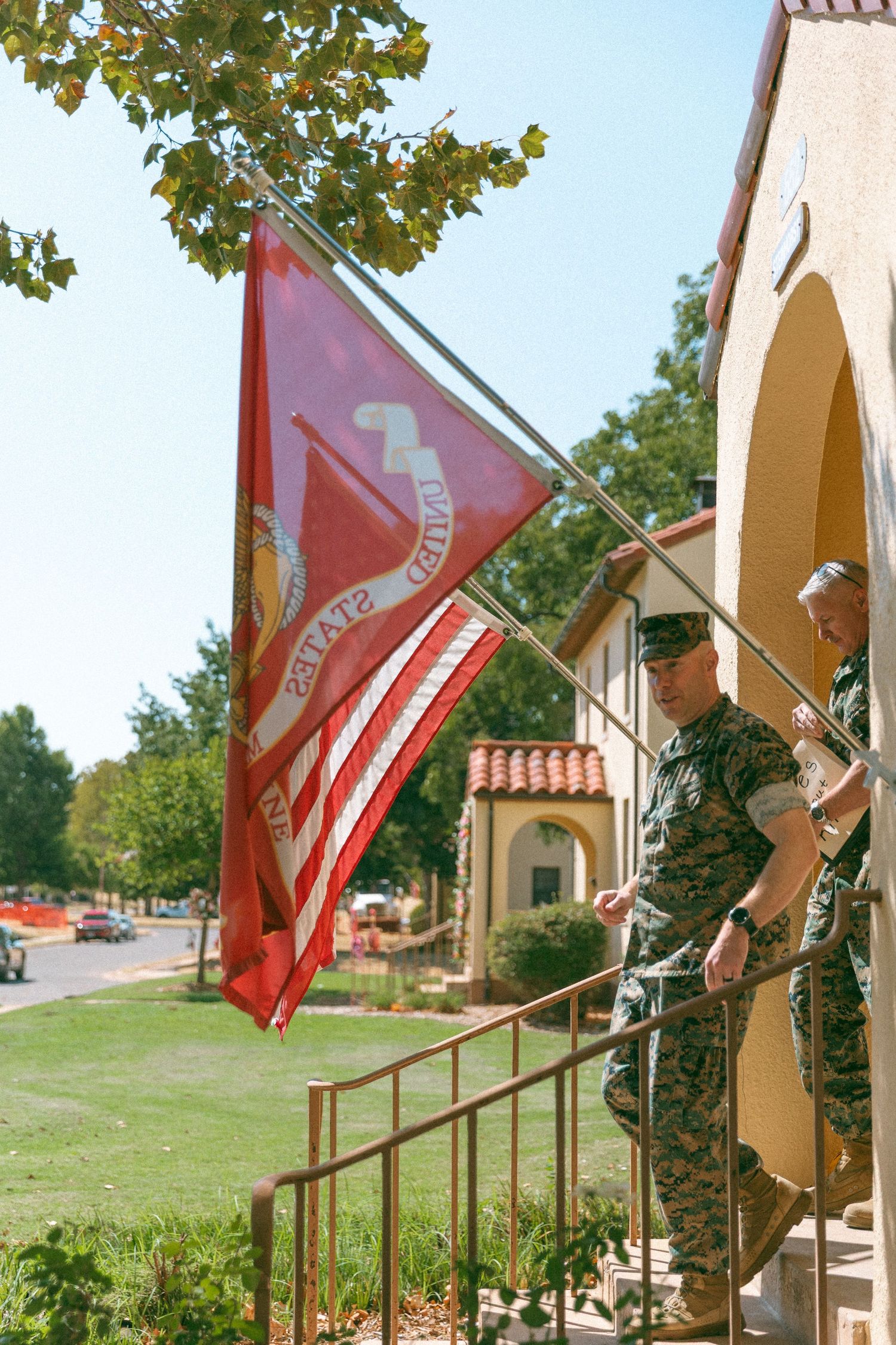 US Marines in camouflage uniforms holding a red United States Marine Corps flag outside a military building.