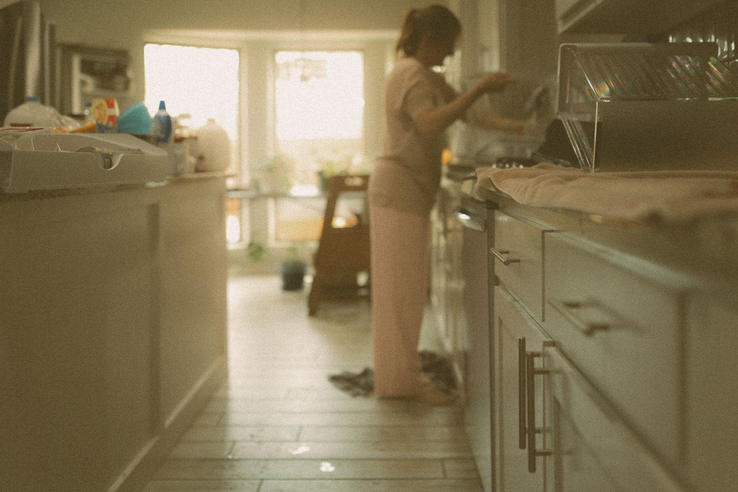 Woman washing dishes in a bright, sunlit kitchen with white cabinets and tiled floors using documentary photographer Liz Perez in Lawton, OK