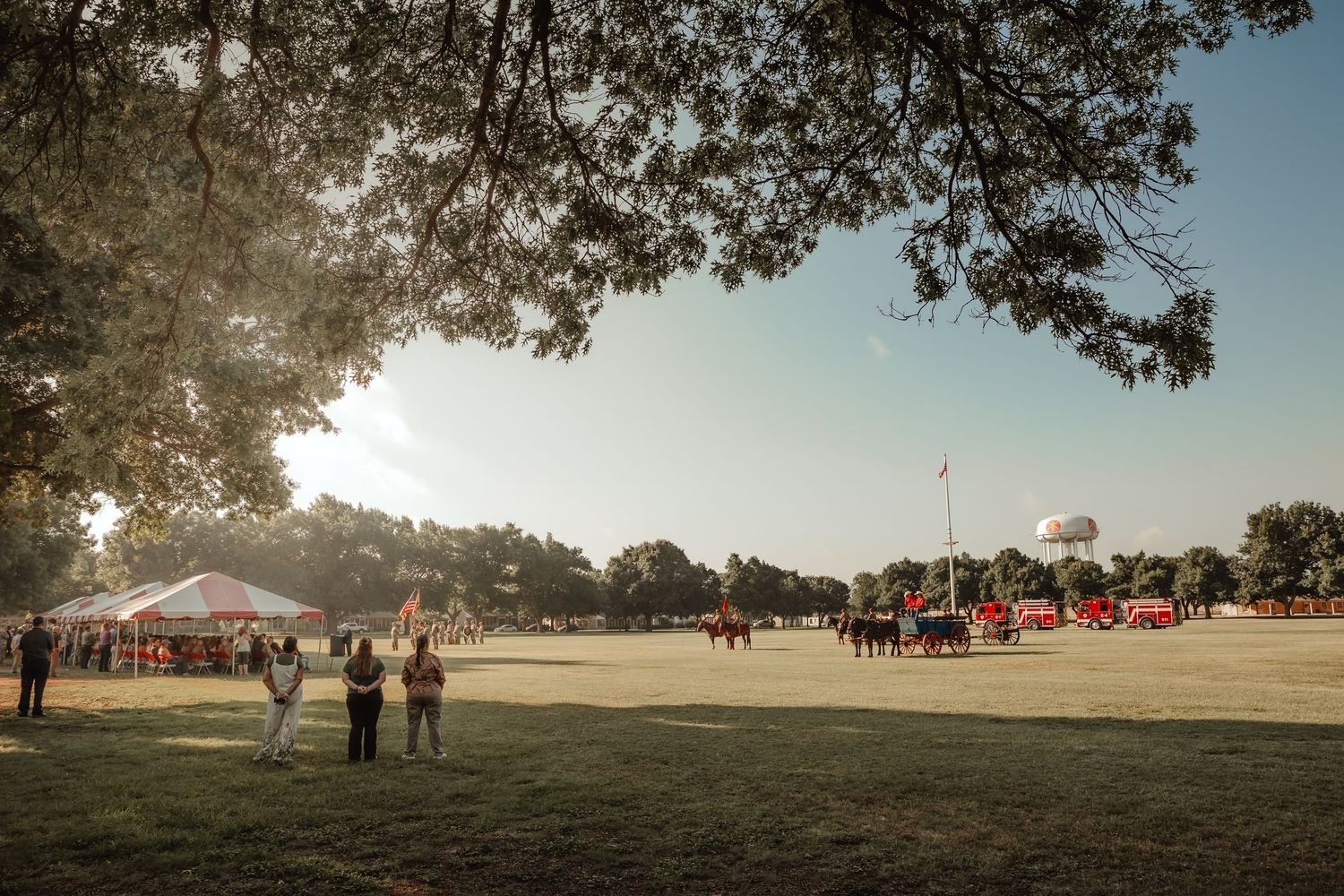 Panoramic view of a grassy field with people gathering under trees at sunset during an outdoor event.