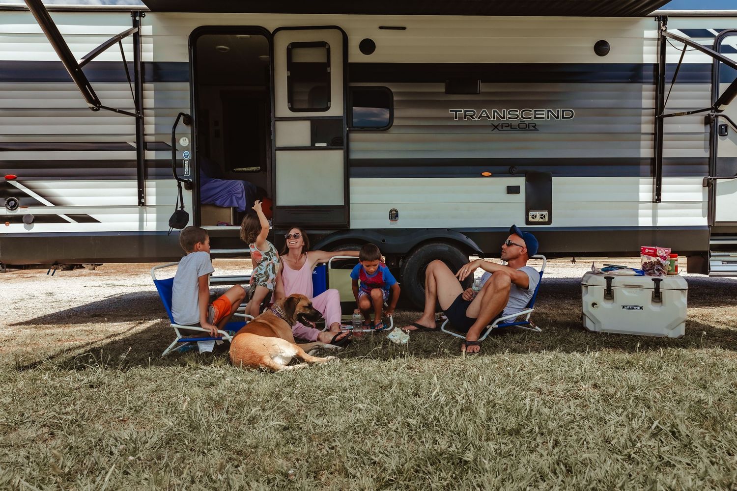 Family relaxing in camping chairs outside a travel trailer RV on a sunny day.