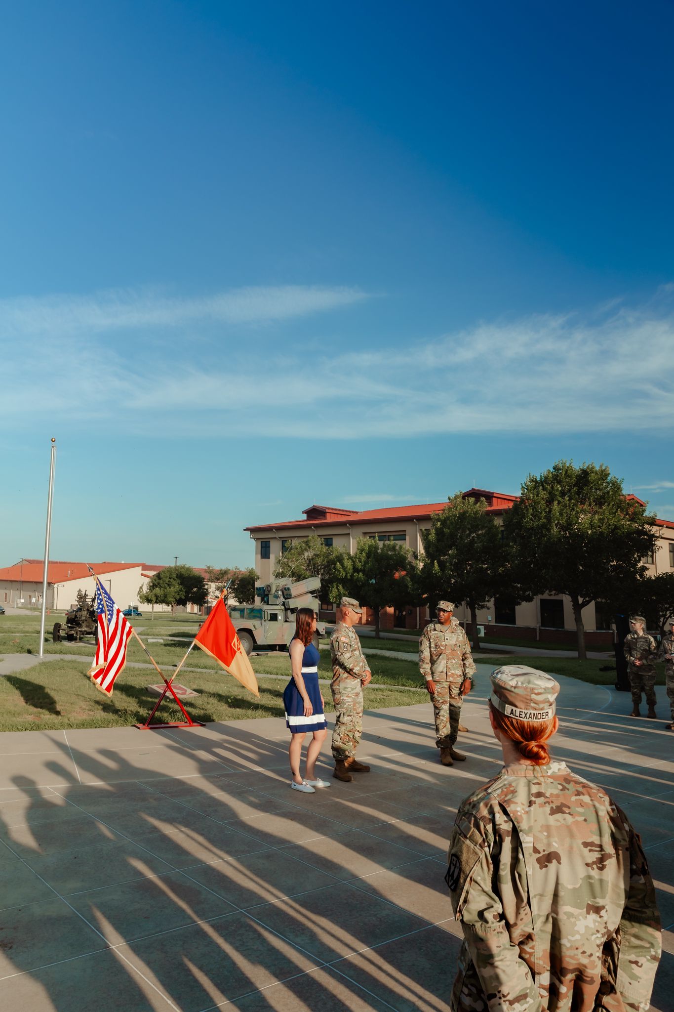 Military personnel in camouflage uniforms participate in a flag ceremony outdoors at a U.S. Army base on a sunny day.