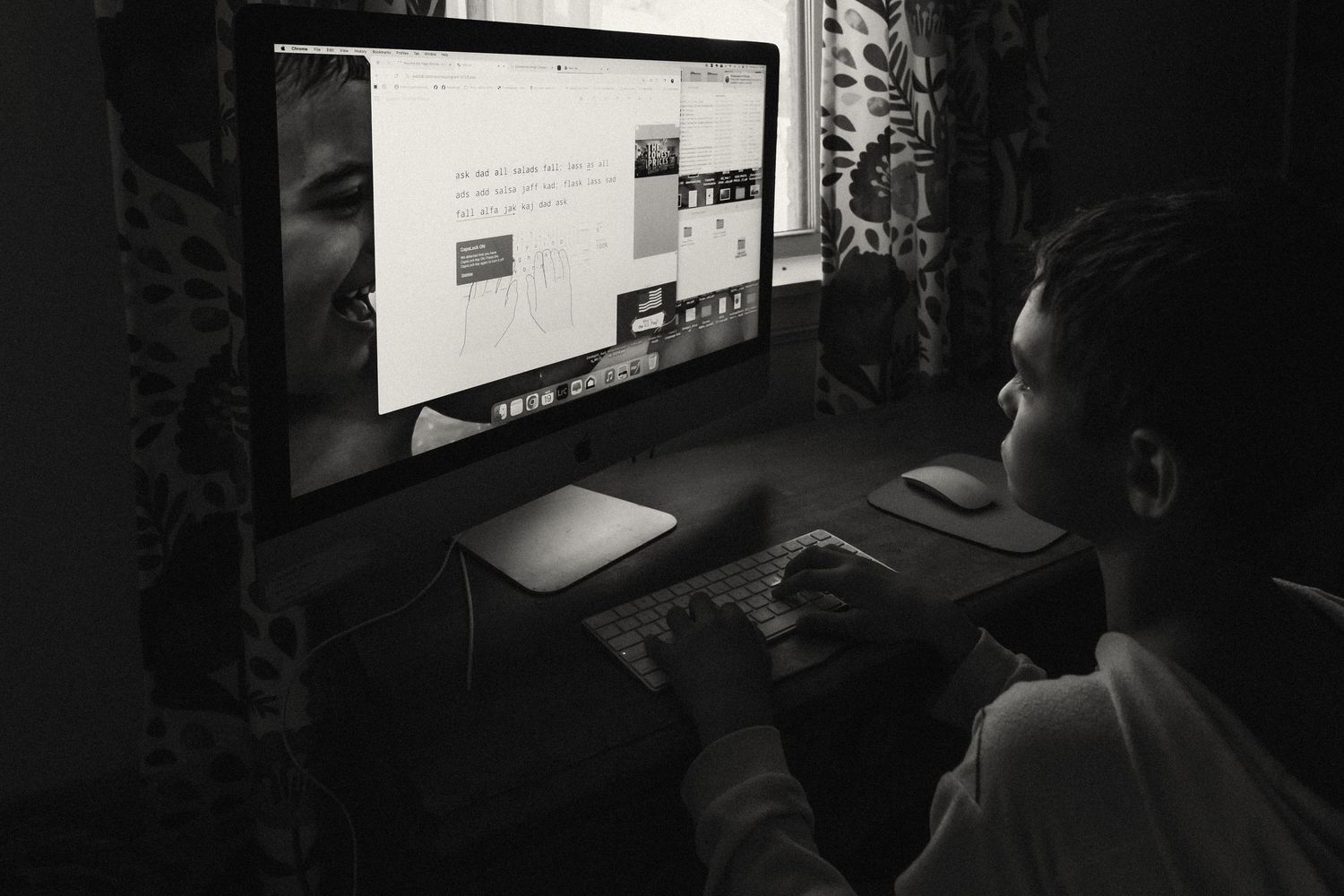 A person works at a computer desk in a dark room with a glowing monitor and keyboard setup.