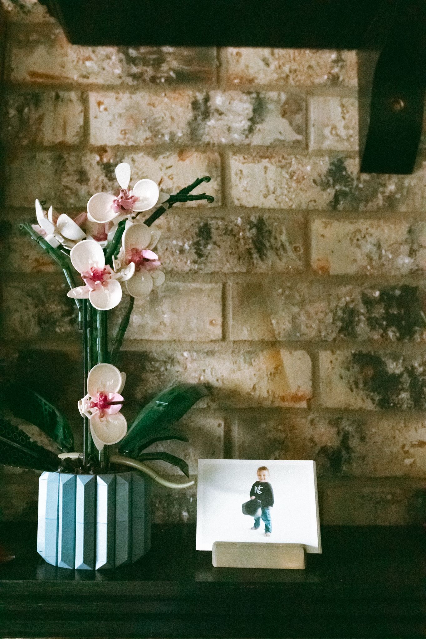 Pink orchids in geometric vase beside a child's photo on wooden shelf against rustic brick wall.