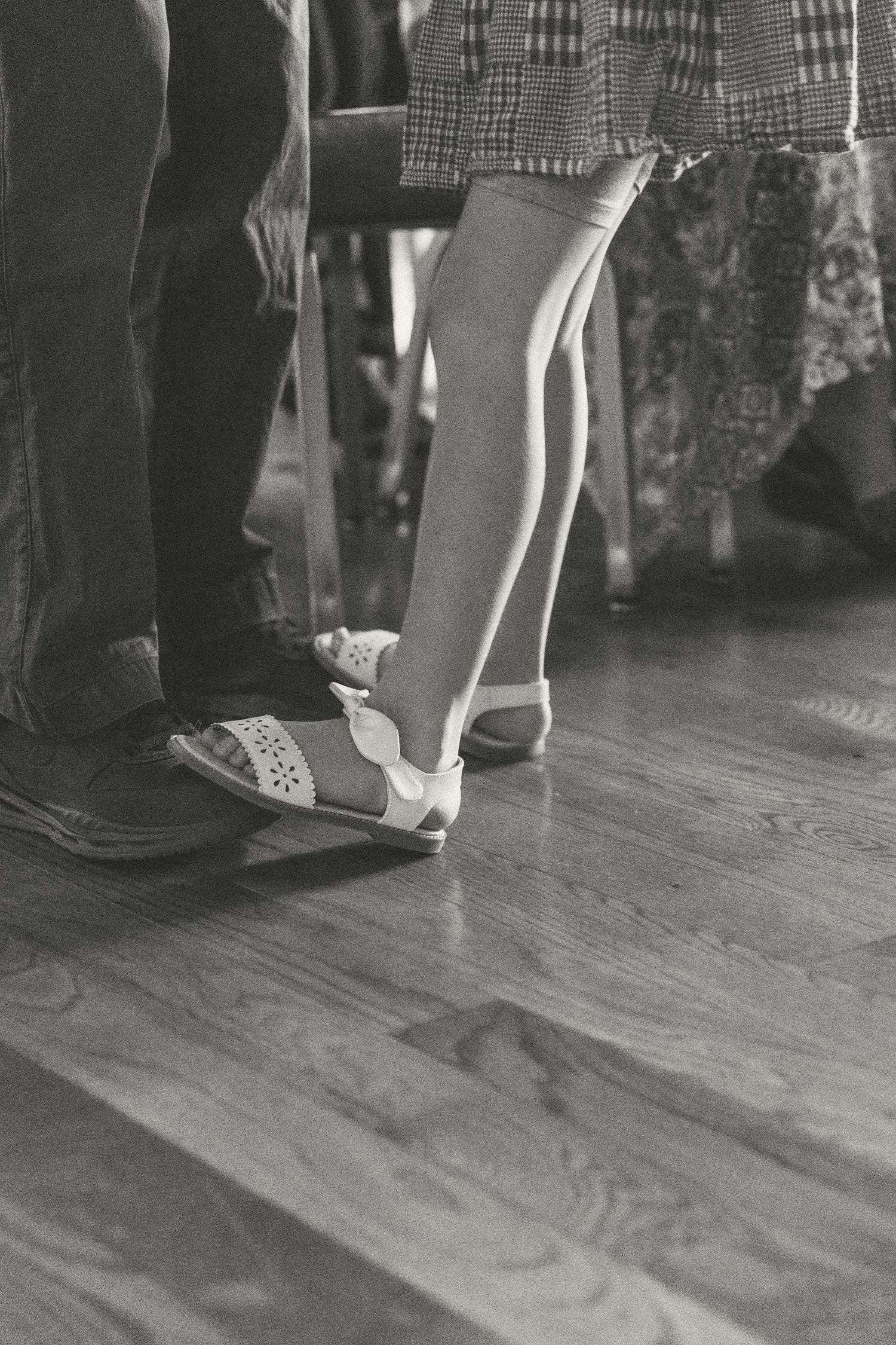 Black and white photo of children and adults feet standing on hardwood floor, girl wearing white sandals.