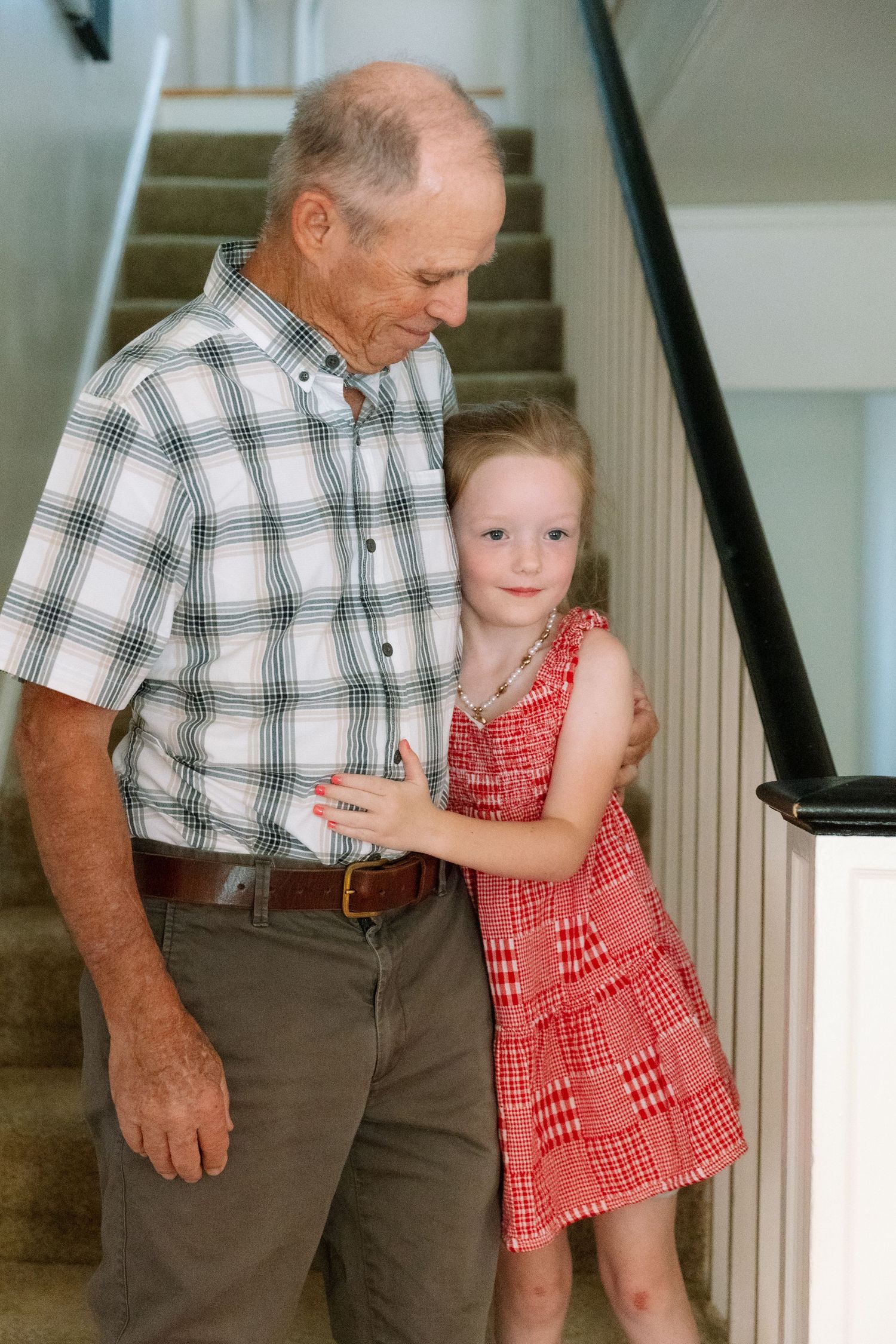 Elderly man in plaid shirt smiling at young girl in red dress standing together by staircase indoors.