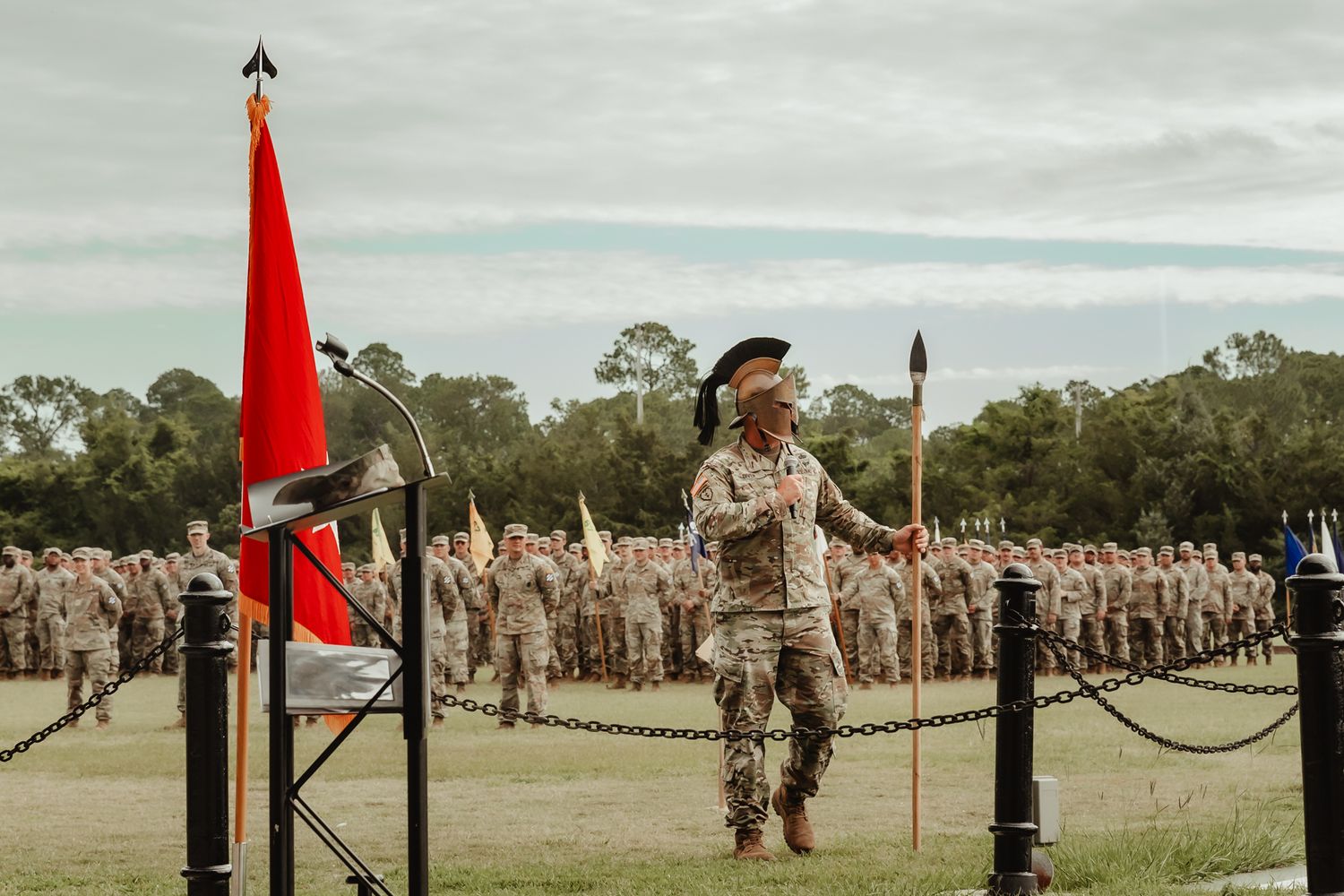 Military personnel stand in formation on a grassy field with red flags visible in the distance.