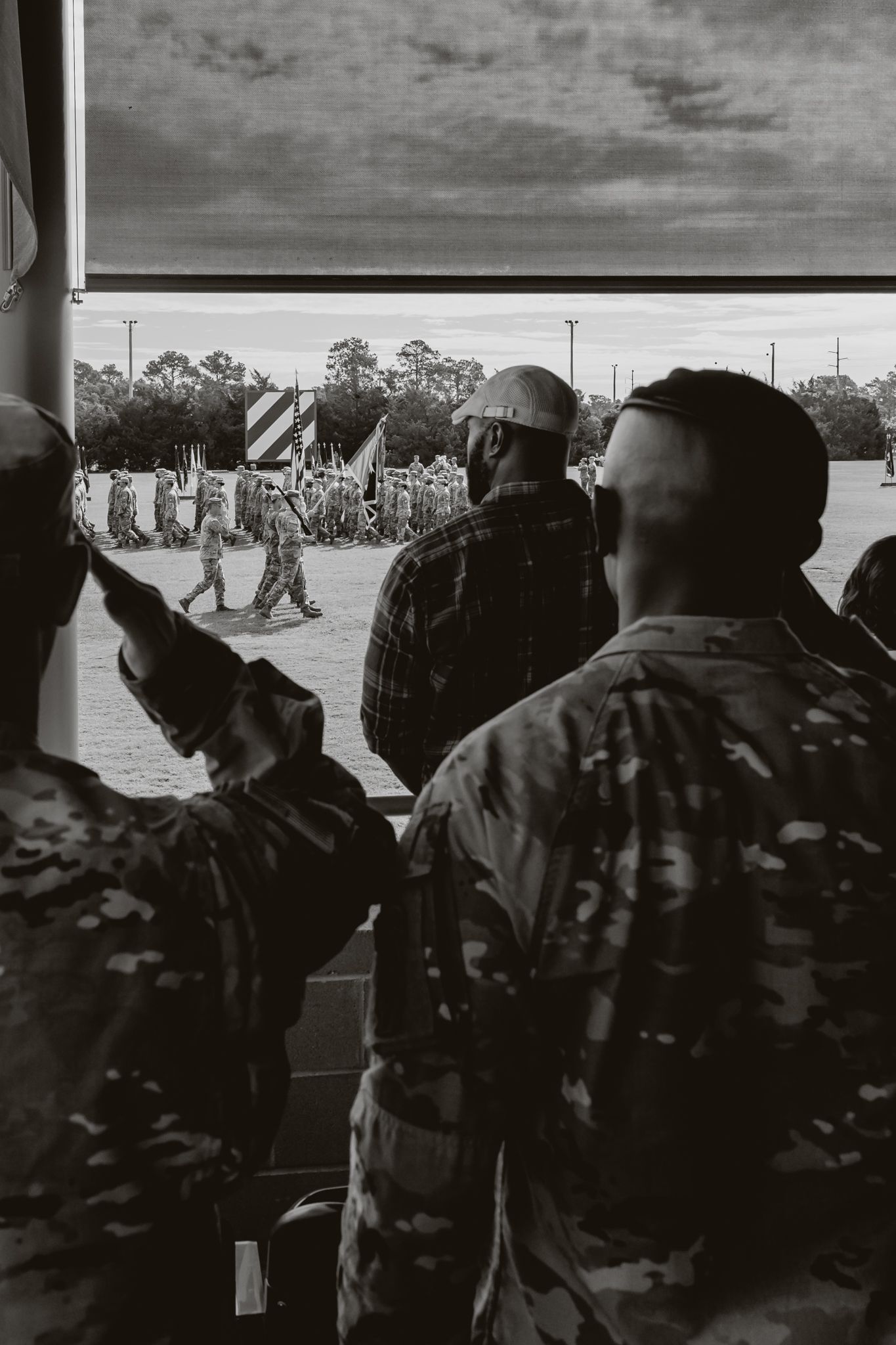 Military personnel in camouflage uniforms observe a formation of soldiers marching in the distance in black and white.