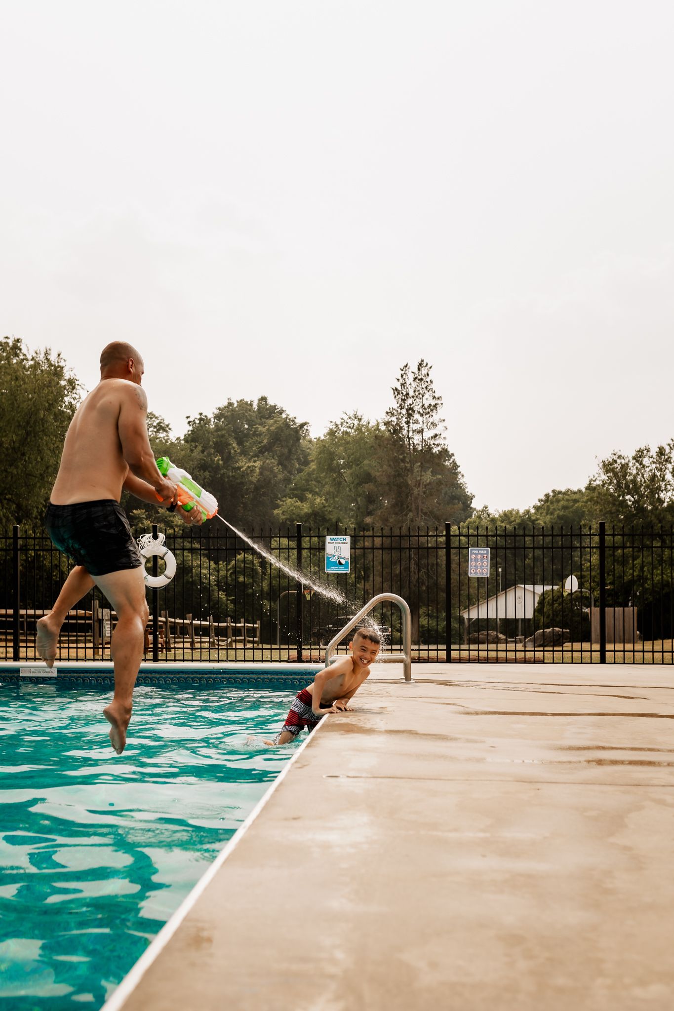 Two people jumping into a swimming pool on a summer day while one holds a pool cleaning net.