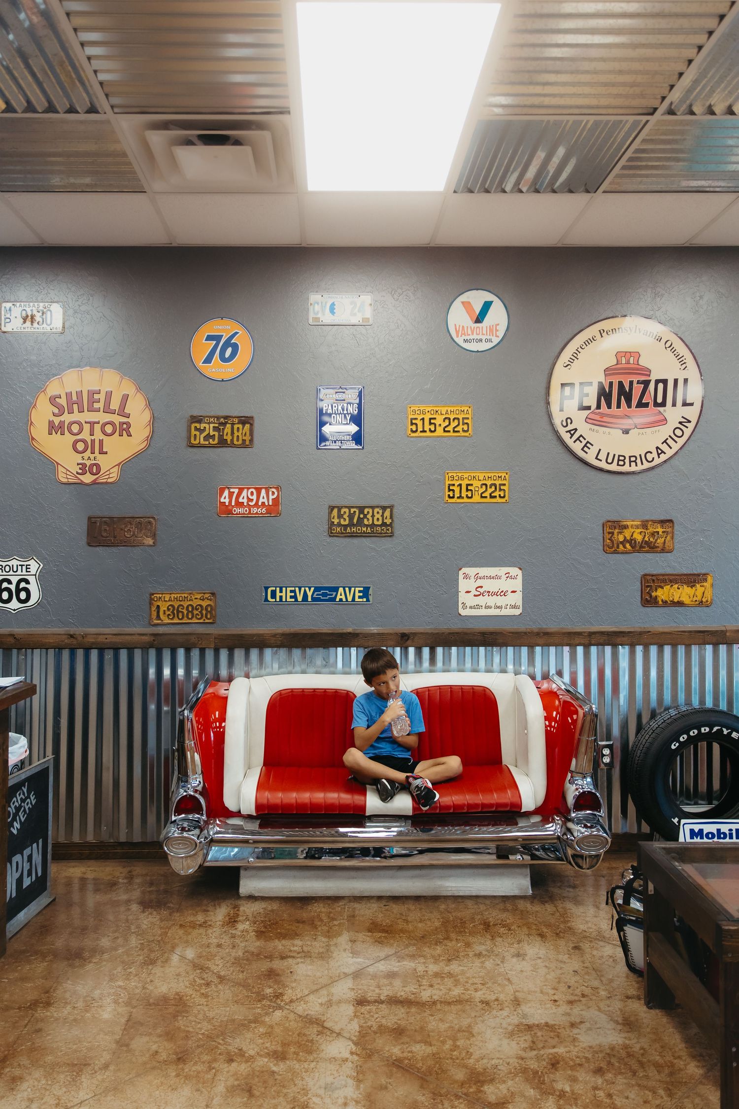 Vintage automotive signs and memorabilia displayed on gray walls above red and white bench seating in a retro-styled space.