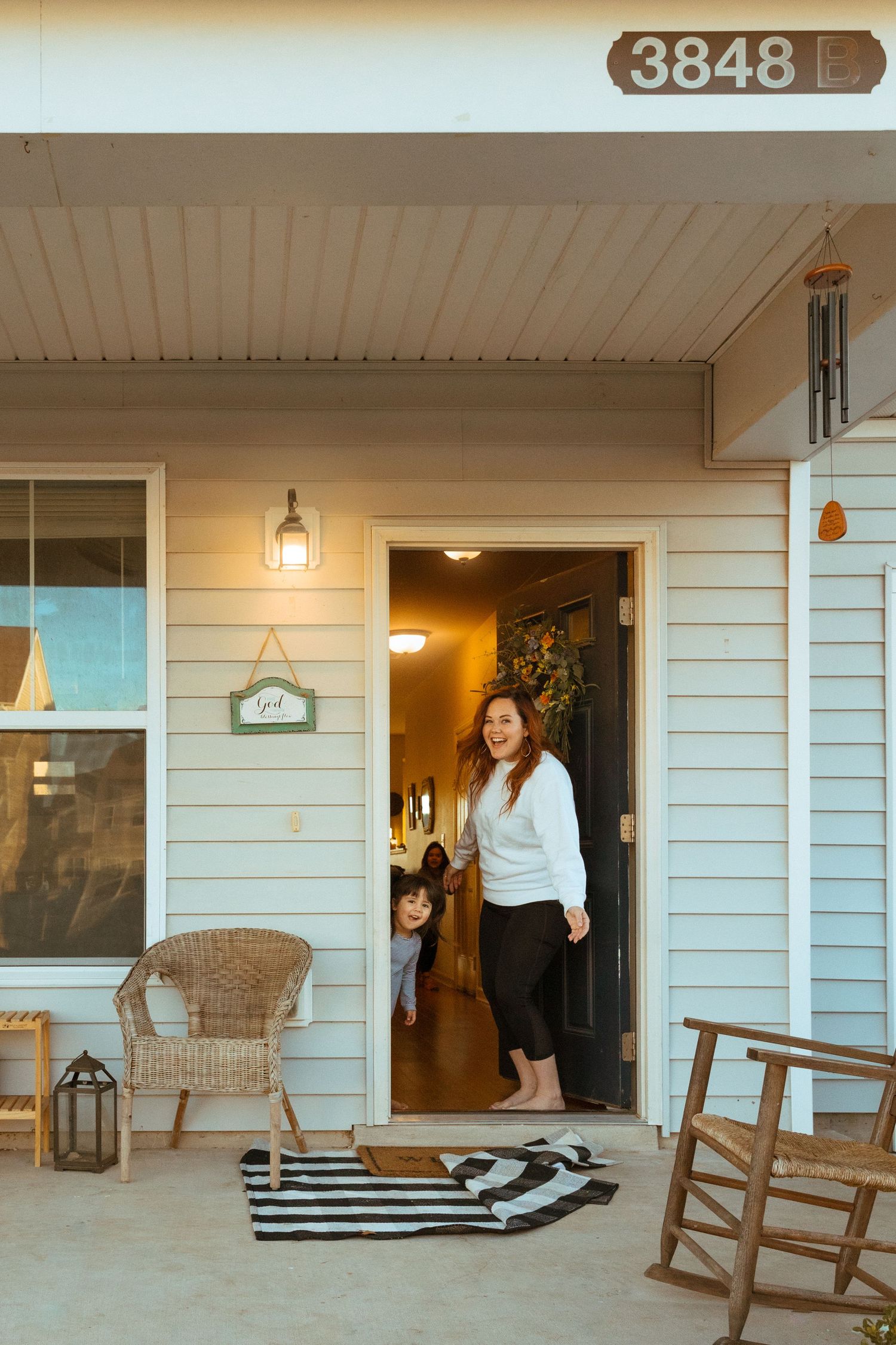 Woman standing in open front door of home numbered 3848 with child behind her and porch furniture visible.