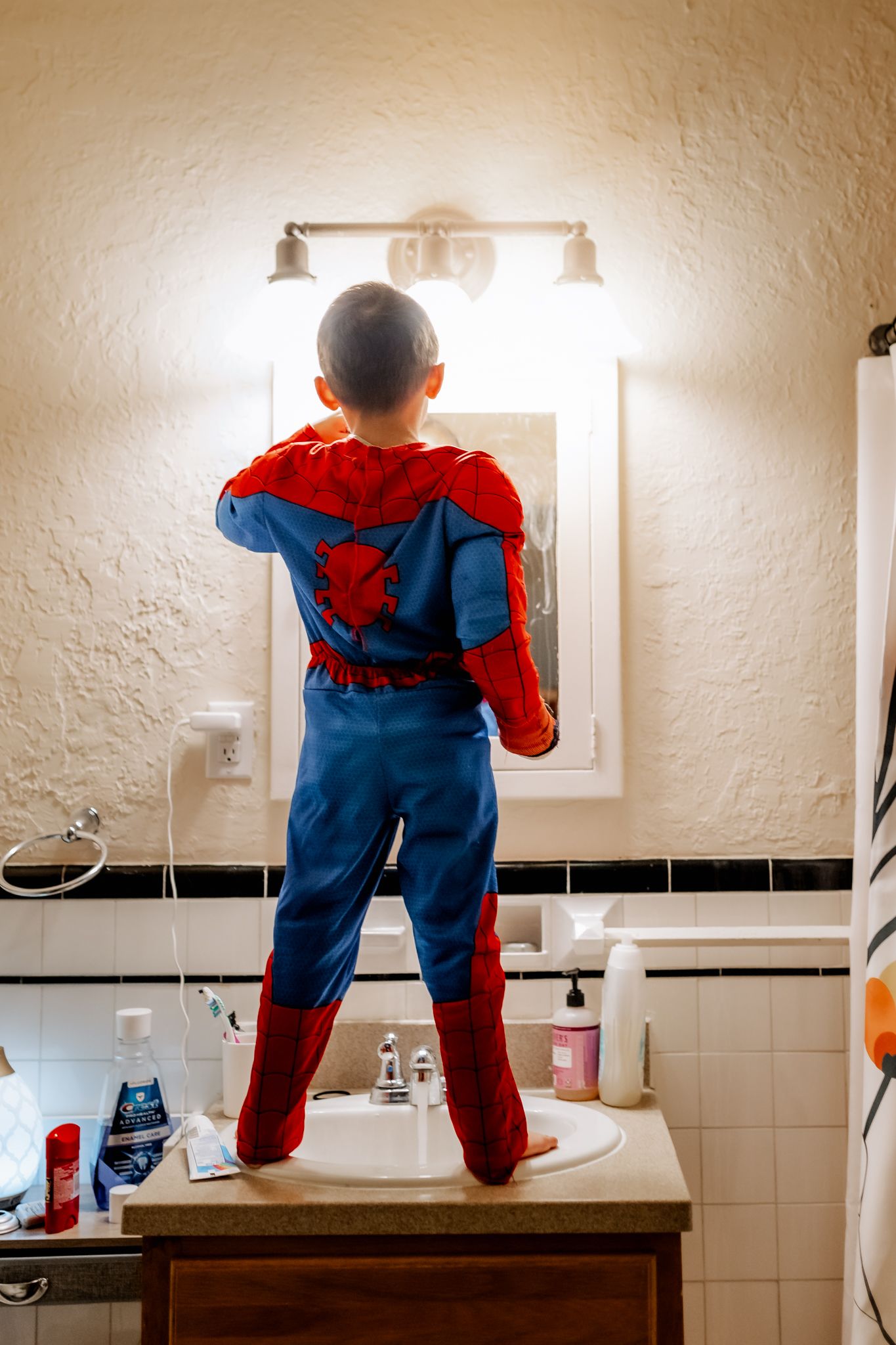 Young boy in Spider-Man costume standing on bathroom counter looking in mirror.