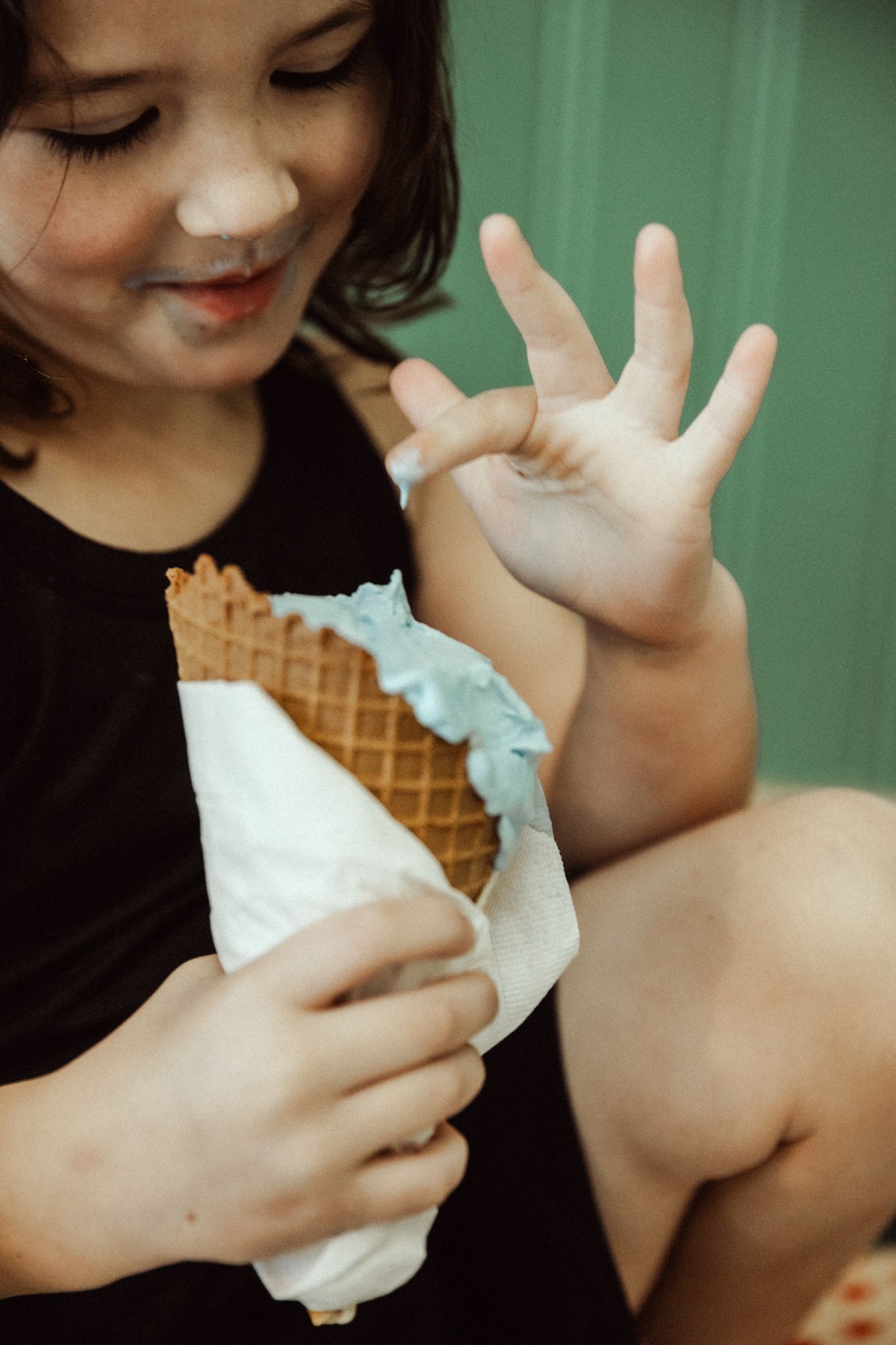 Young girl in black shirt smiling while holding a melting blue ice cream waffle cone with sticky hands.